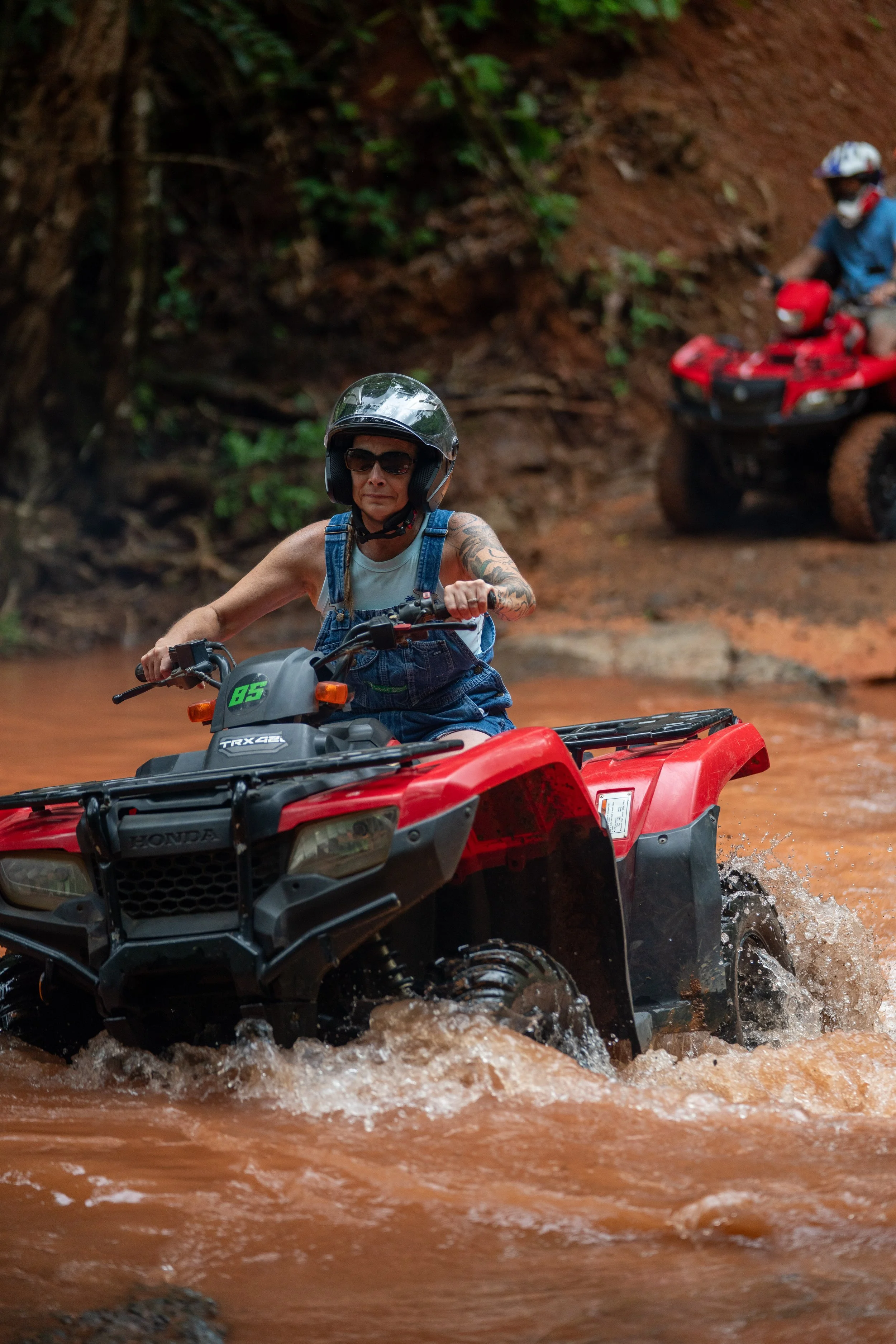A woman wearing sunglasses and a helmet riding a red all-terrain vehicle through a muddy trail, with a man in the background on another ATV.