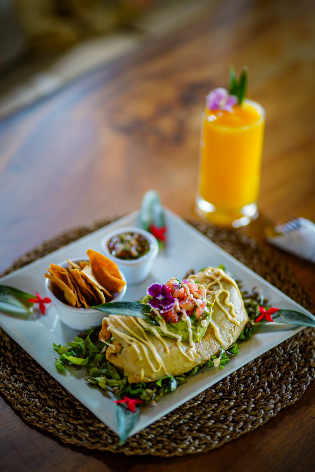 Mexican dish with tacos topped with diced tomatoes, onions, and cilantro, served with sides of salsas and chips, and a glass of orange juice with a flower garnish in the background.