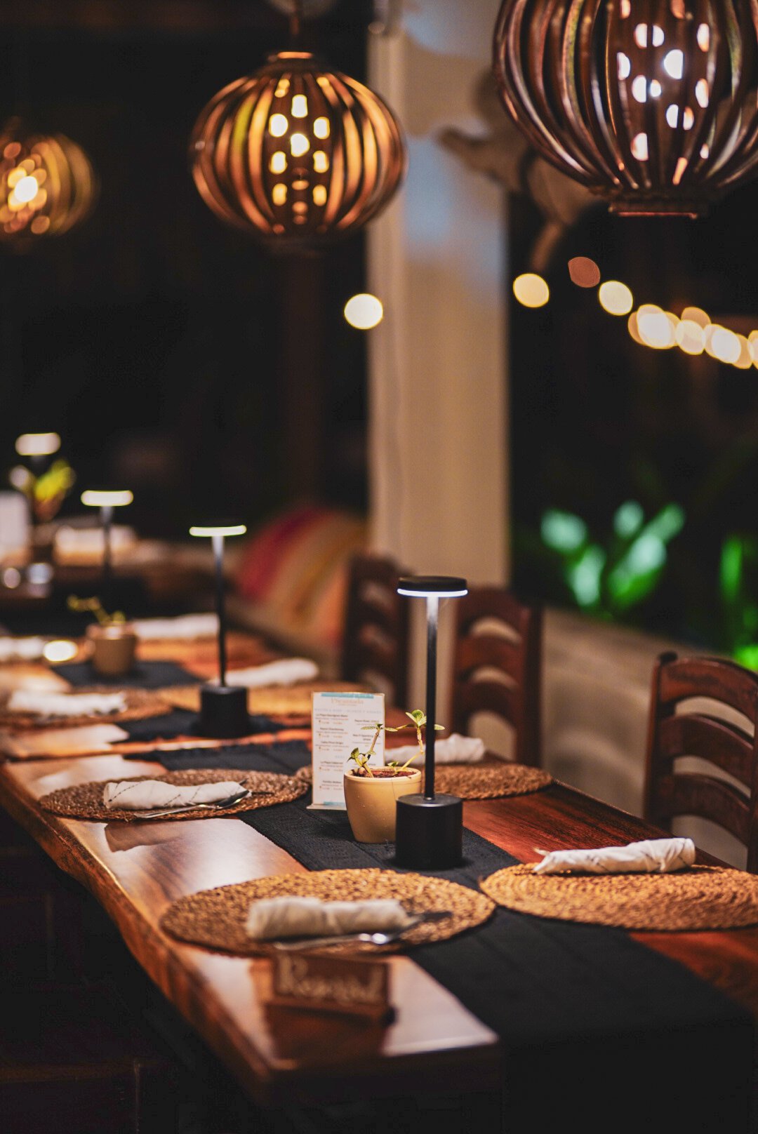 A dimly lit restaurant table set for dining, featuring woven placemats, rolled napkins, small table lamps, and decorative hanging lantern lights.