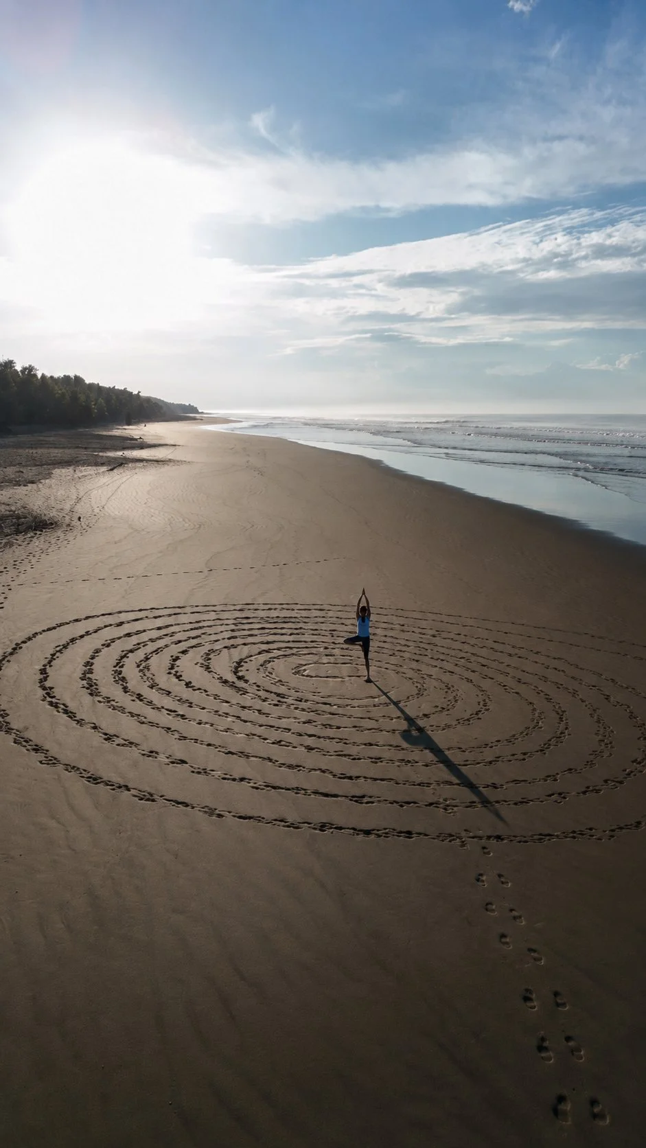 A person practicing yoga on a sandy beach, with concentric circles drawn in the sand around them and the ocean waves in the background.
