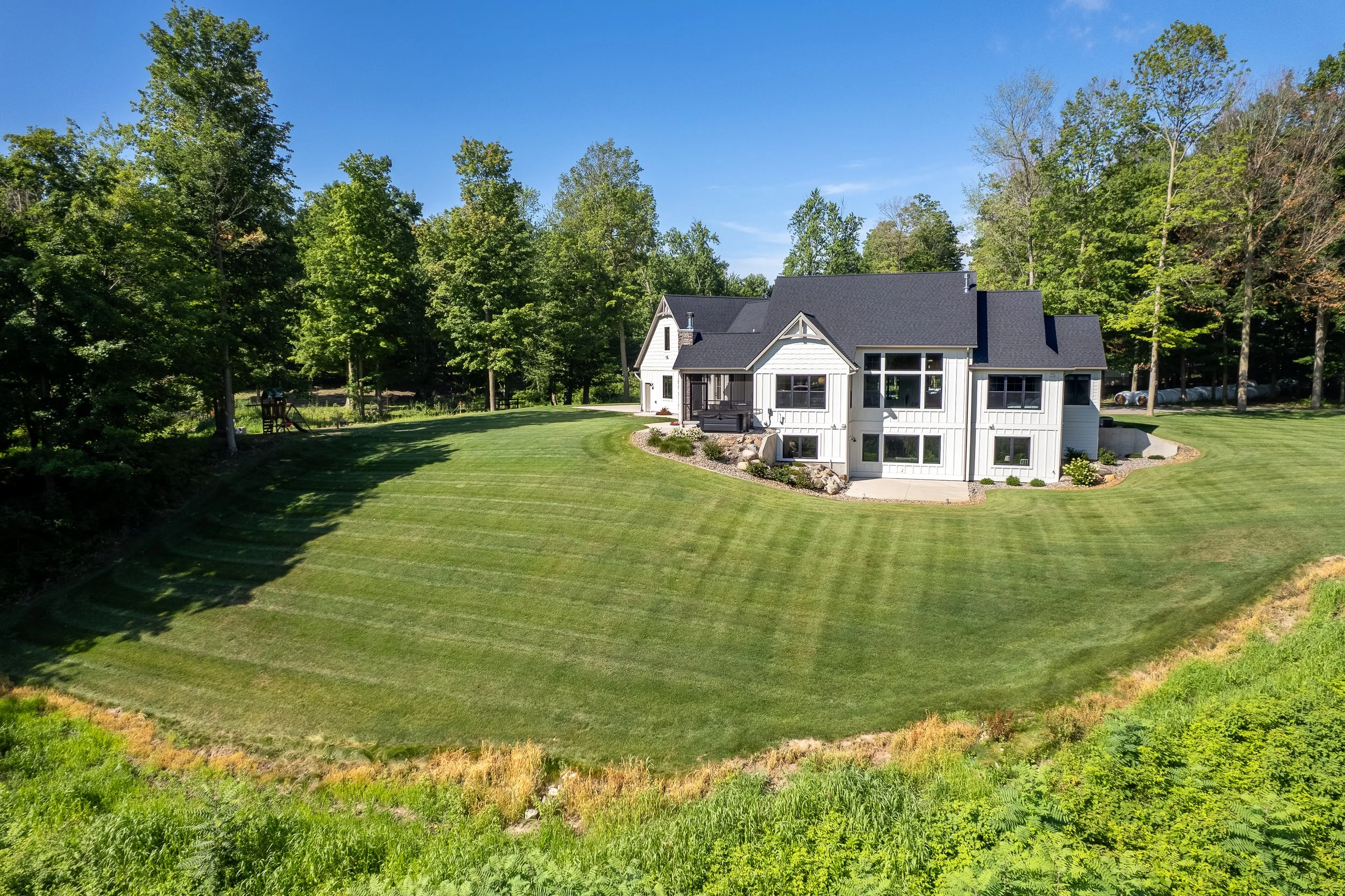 A large white house with black roof shingles sits on a spacious green lawn bordered by trees under a clear blue sky.