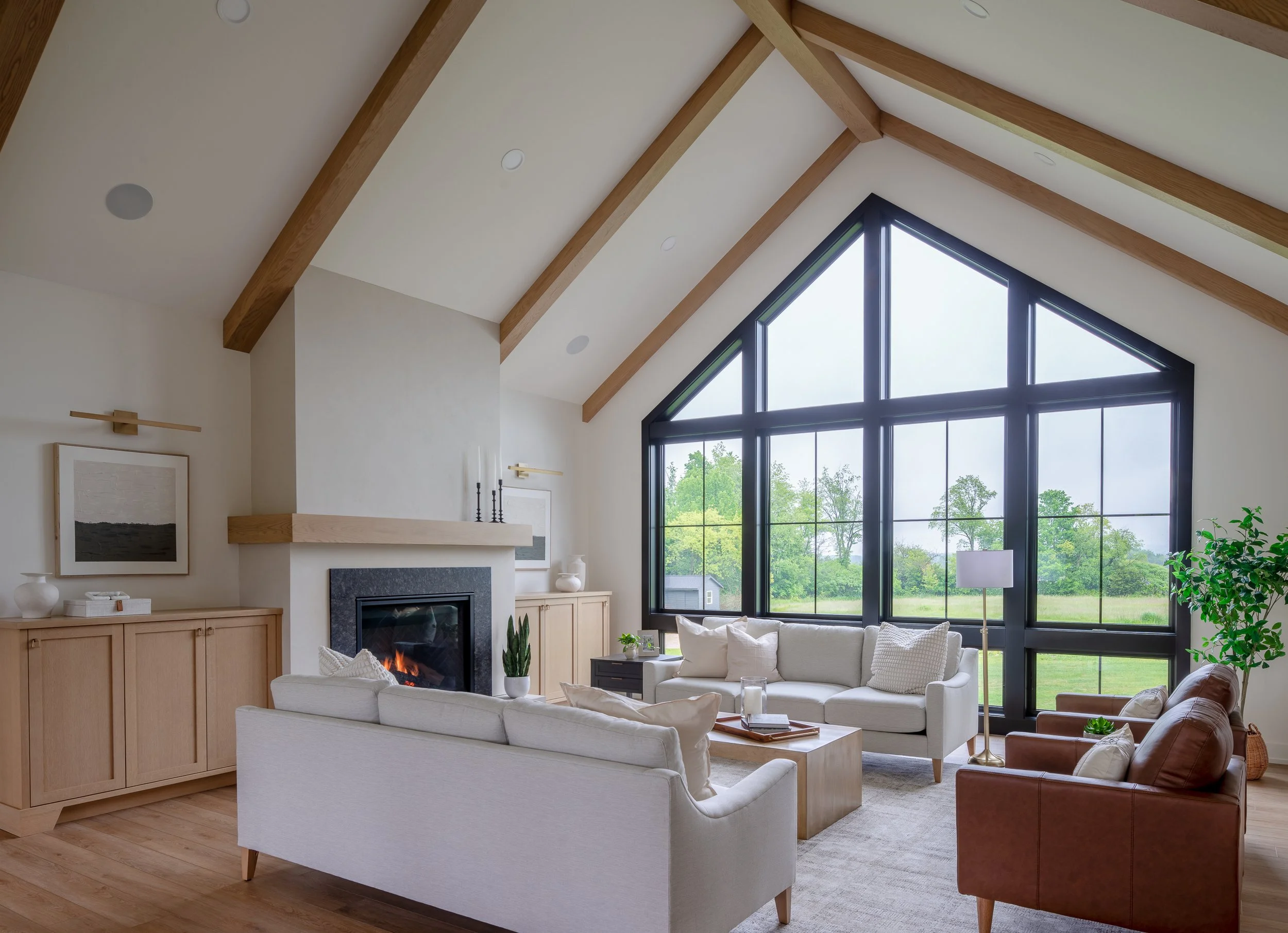 Living room with a large window, white and brown couches, a fireplace, wooden furniture, and a view of green trees outside.