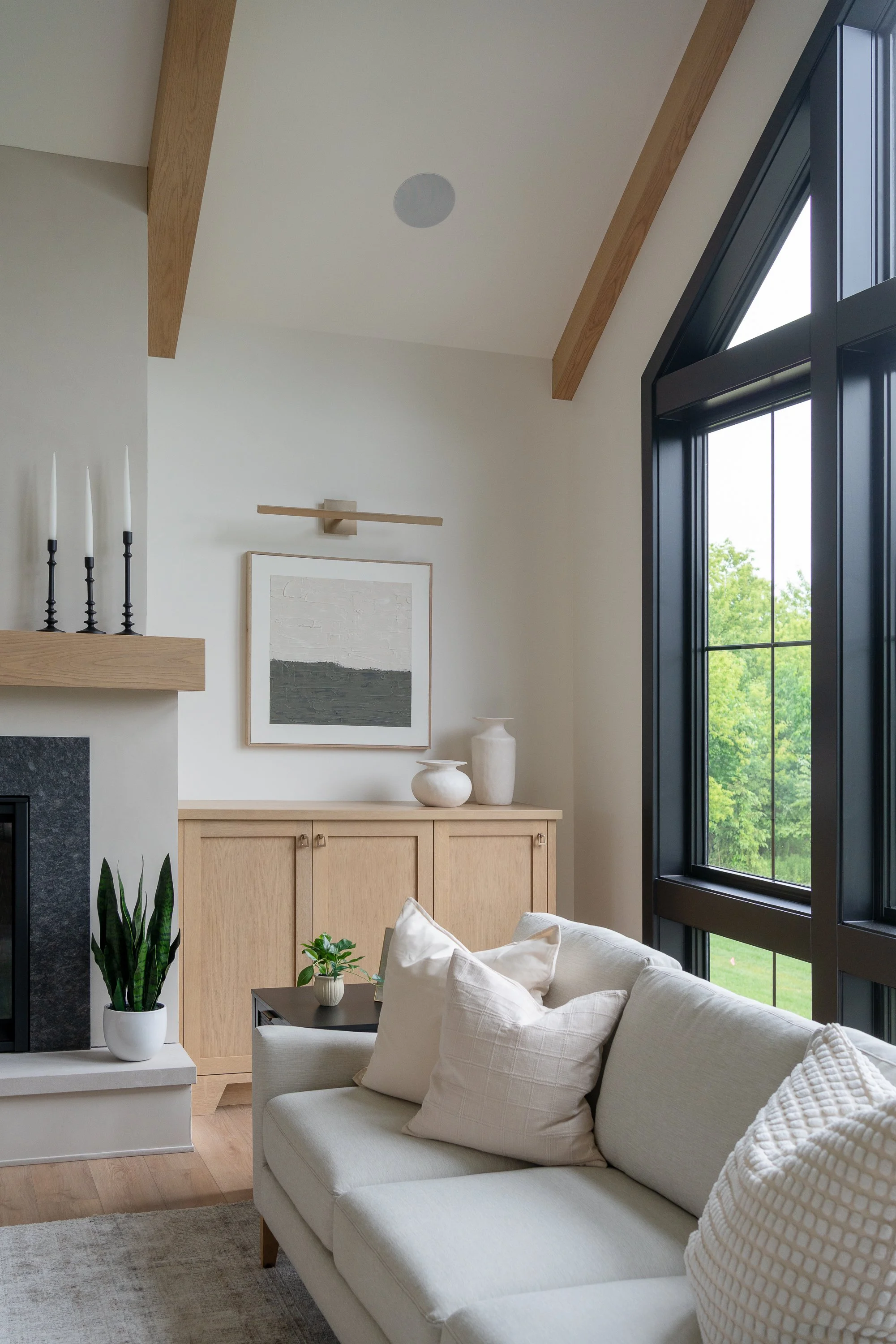 Living room with beige sofa, throw pillows, black window frames, wooden cabinet, white vases, artwork, and fireplace with potted plant.