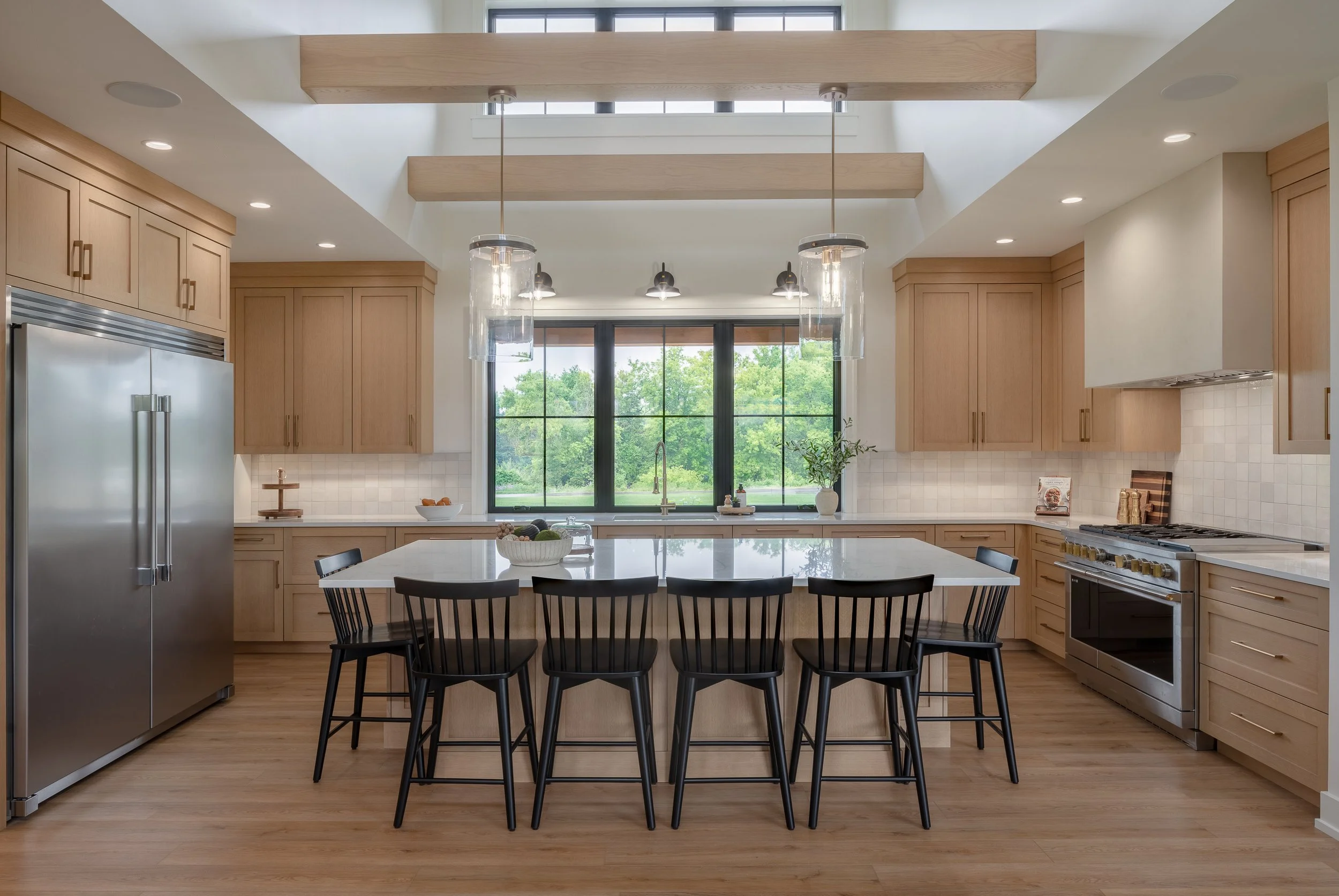 Modern kitchen with light wood cabinetry, large white island, black chairs, stainless steel refrigerator, and a window overlooking greenery.