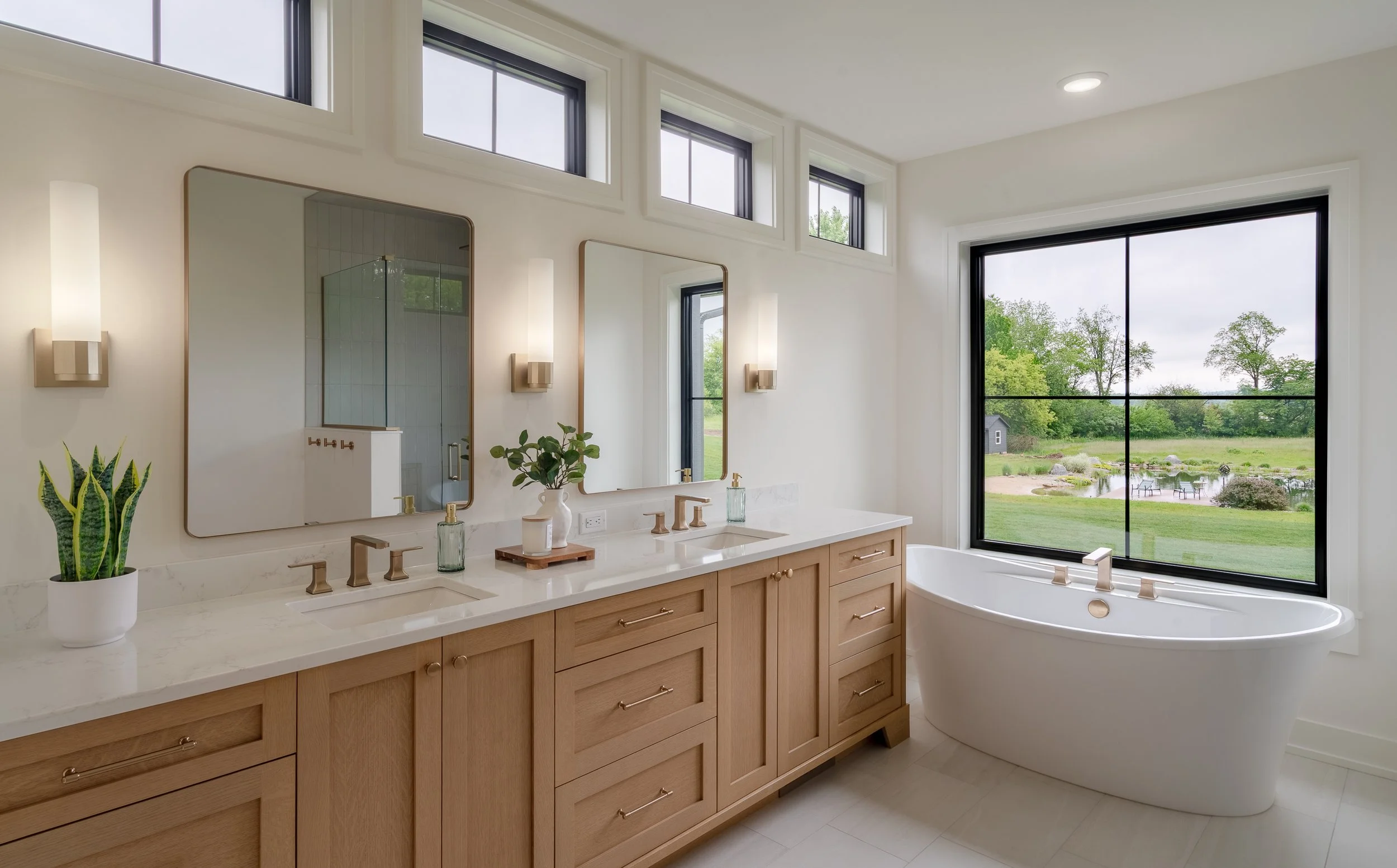 Modern bathroom with double vanity, large mirror, potted plant, and a bathtub by a large window overlooking a green landscape with trees and a pond.