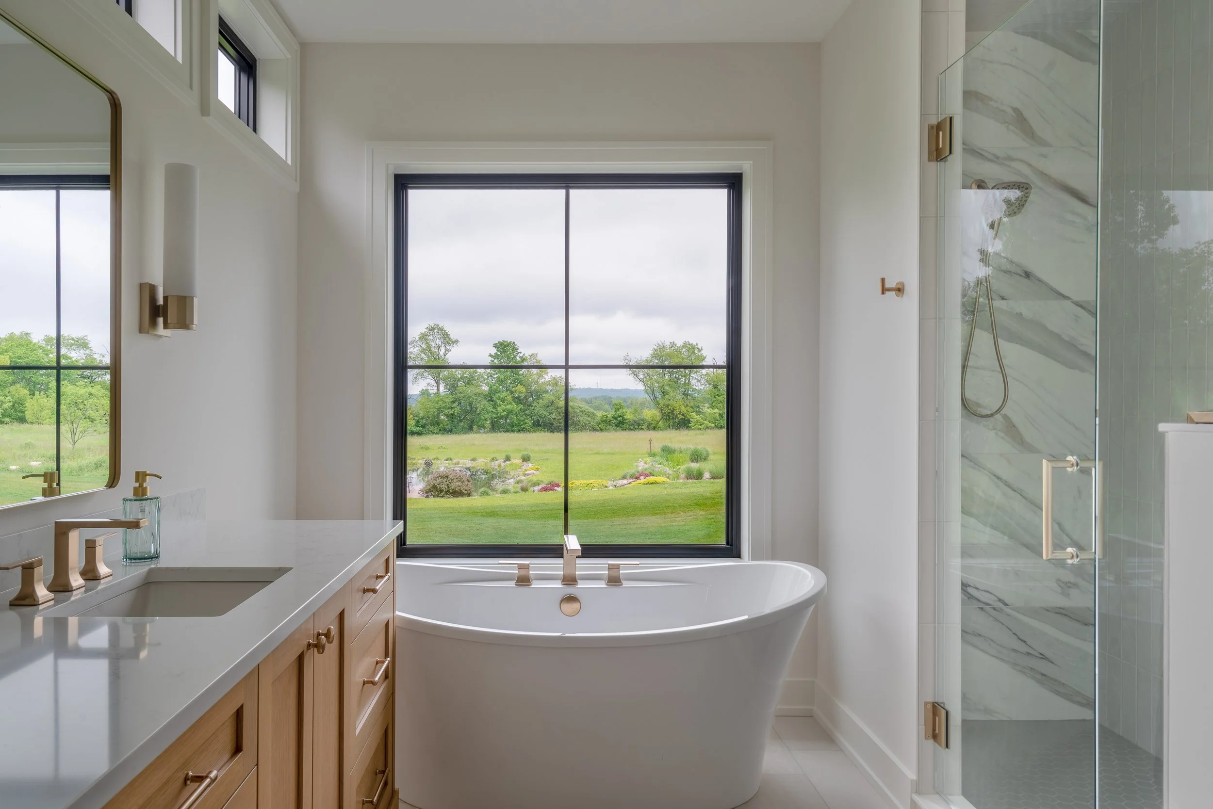 Modern bathroom with a freestanding bathtub near a large window overlooking green trees and a lawn, with a walk-in shower with a glass door on the right side and a wooden vanity with a sink on the left.
