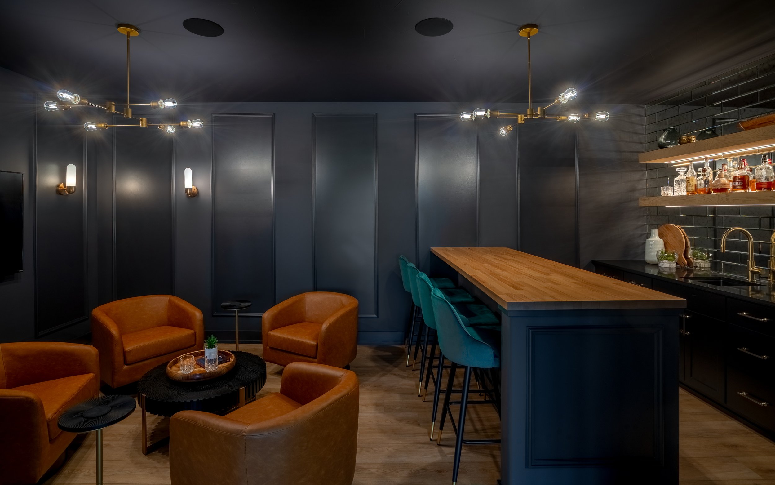 Cozy bar interior with dark walls, modern lighting fixtures, leather armchairs, a wooden bar counter with teal barstools, and shelves with bottles of alcohol.
