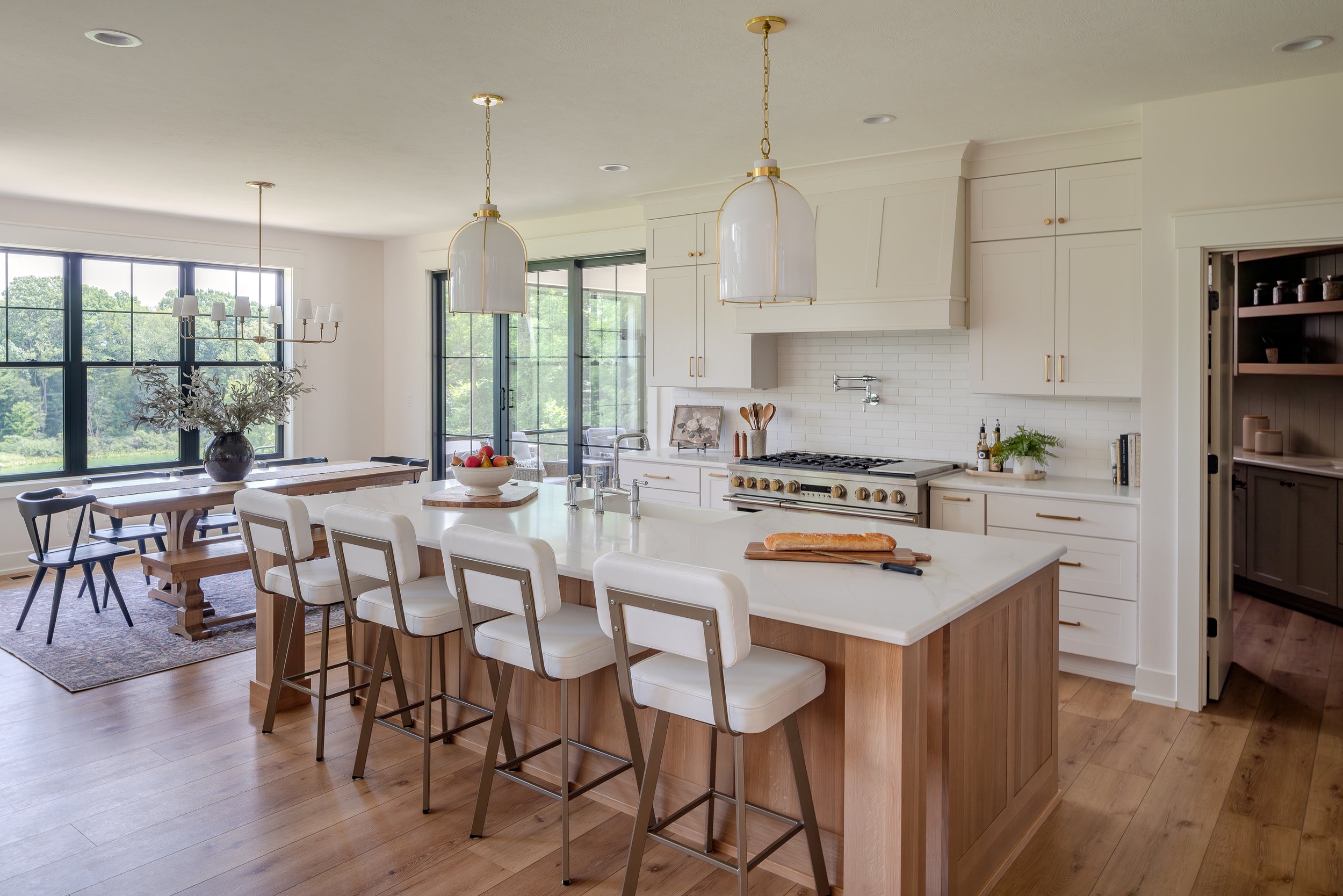 Bright kitchen with white cabinets, a large island with white countertop and four bar stools, a stove, and a dining area with a wooden table and black chairs by large windows with a view of greenery.