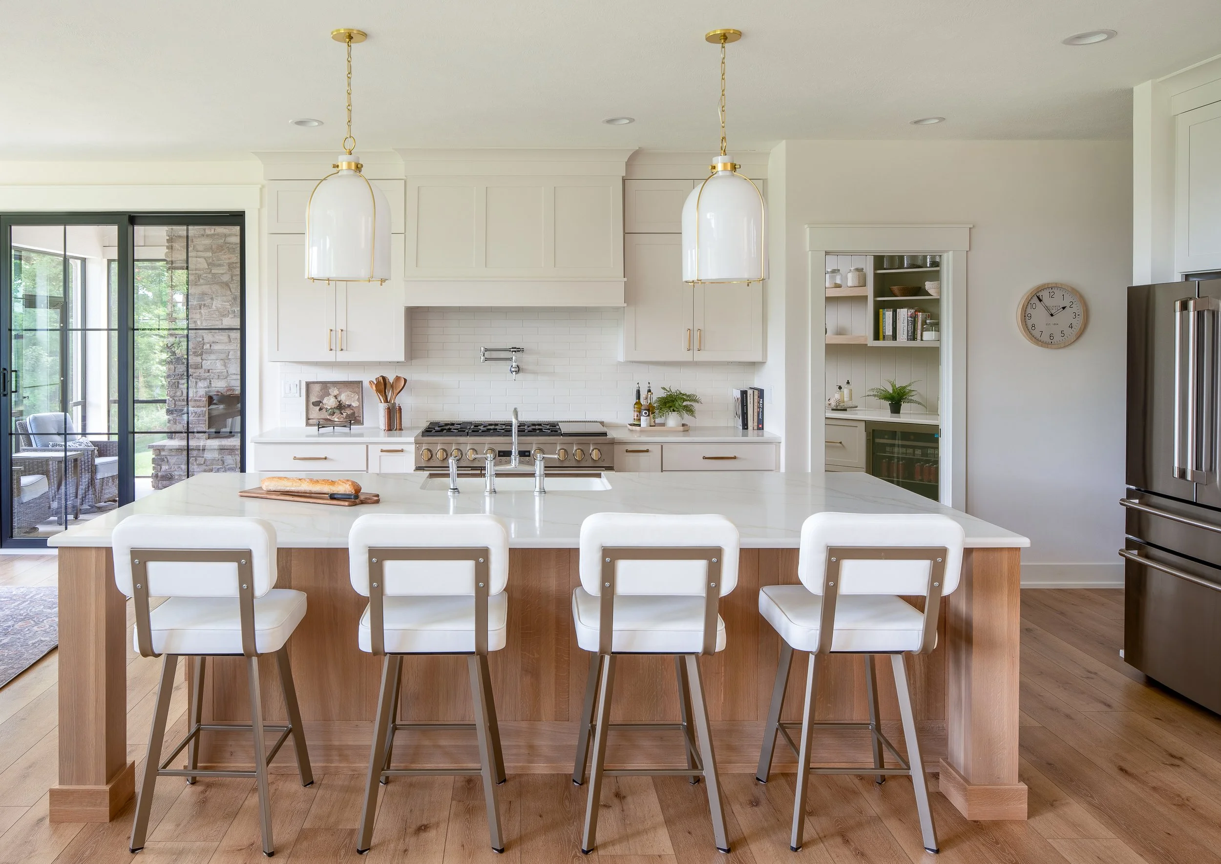 Modern kitchen with white cabinetry, a large white island with a wood base, four white bar stools, pendant lights, a stainless steel refrigerator, and a sliding glass door leading to an outdoor seating area.