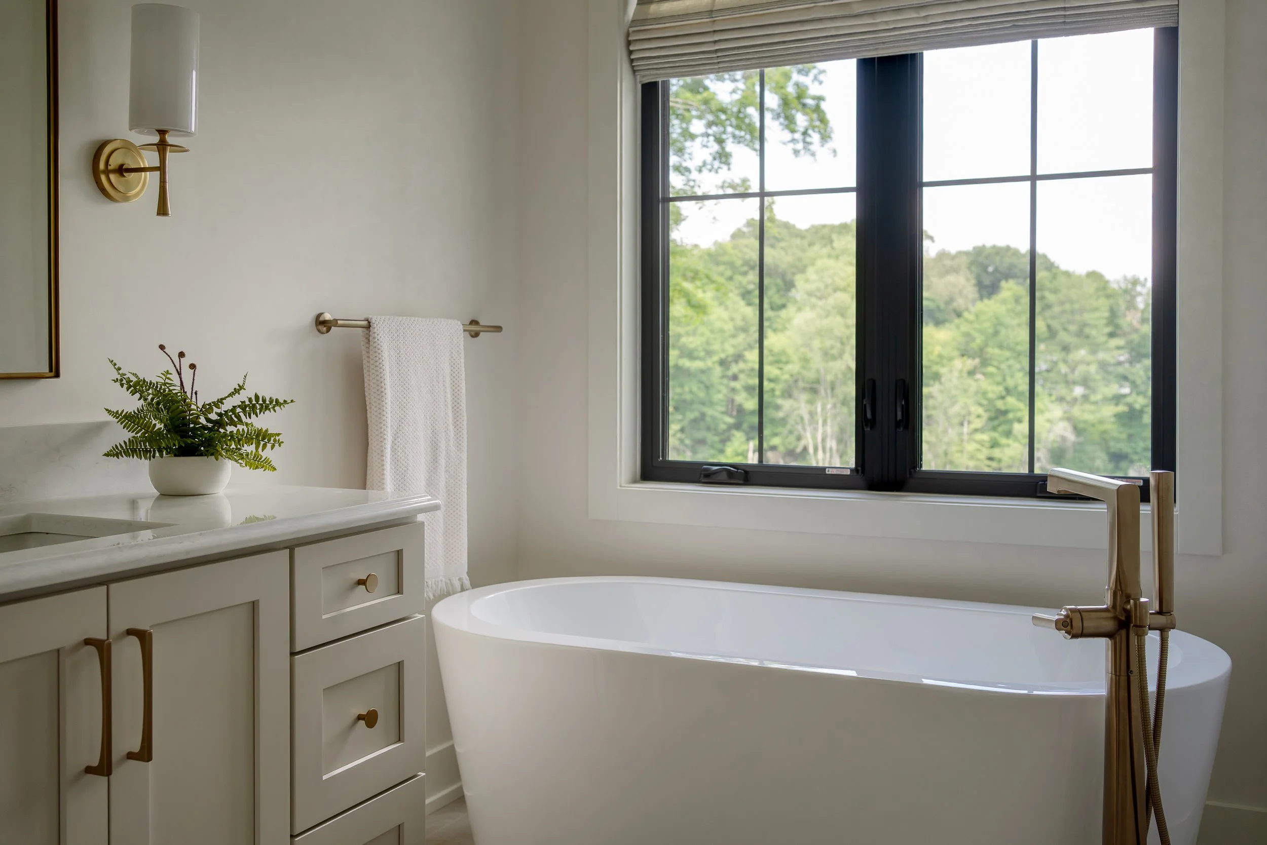 Bathroom with a white bathtub in front of a large window showing a green landscape, a white vanity with a plant, and a wall-mounted light fixture.