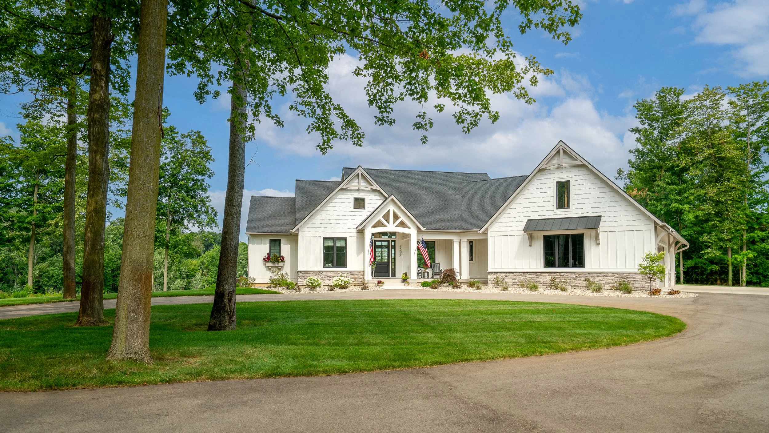 White modern house with black roof, front porch, and landscaping, surrounded by trees on a sunny day.