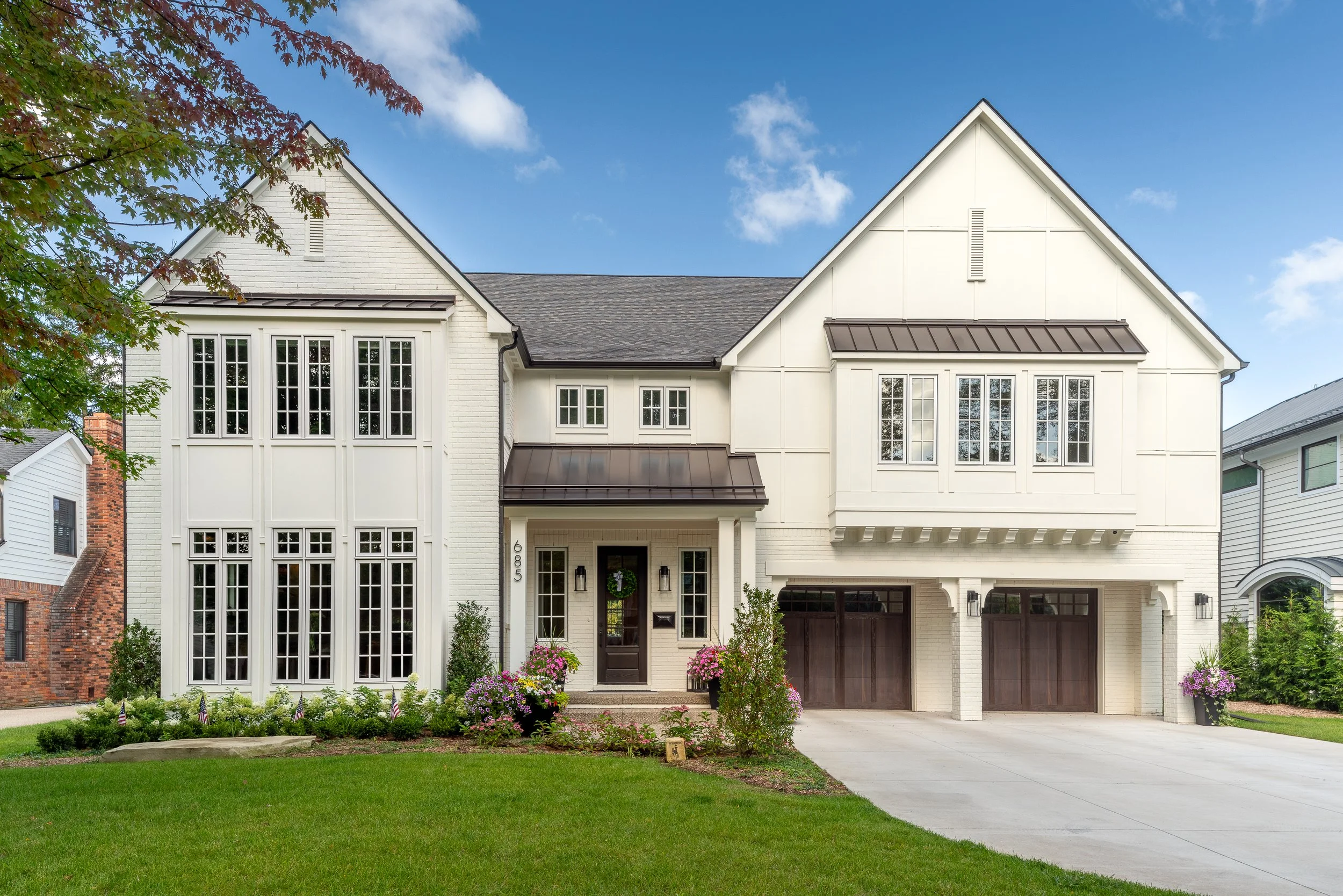 Modern two-story white house with large windows, a driveway, and a well-maintained front yard with flowering plants and a green lawn.