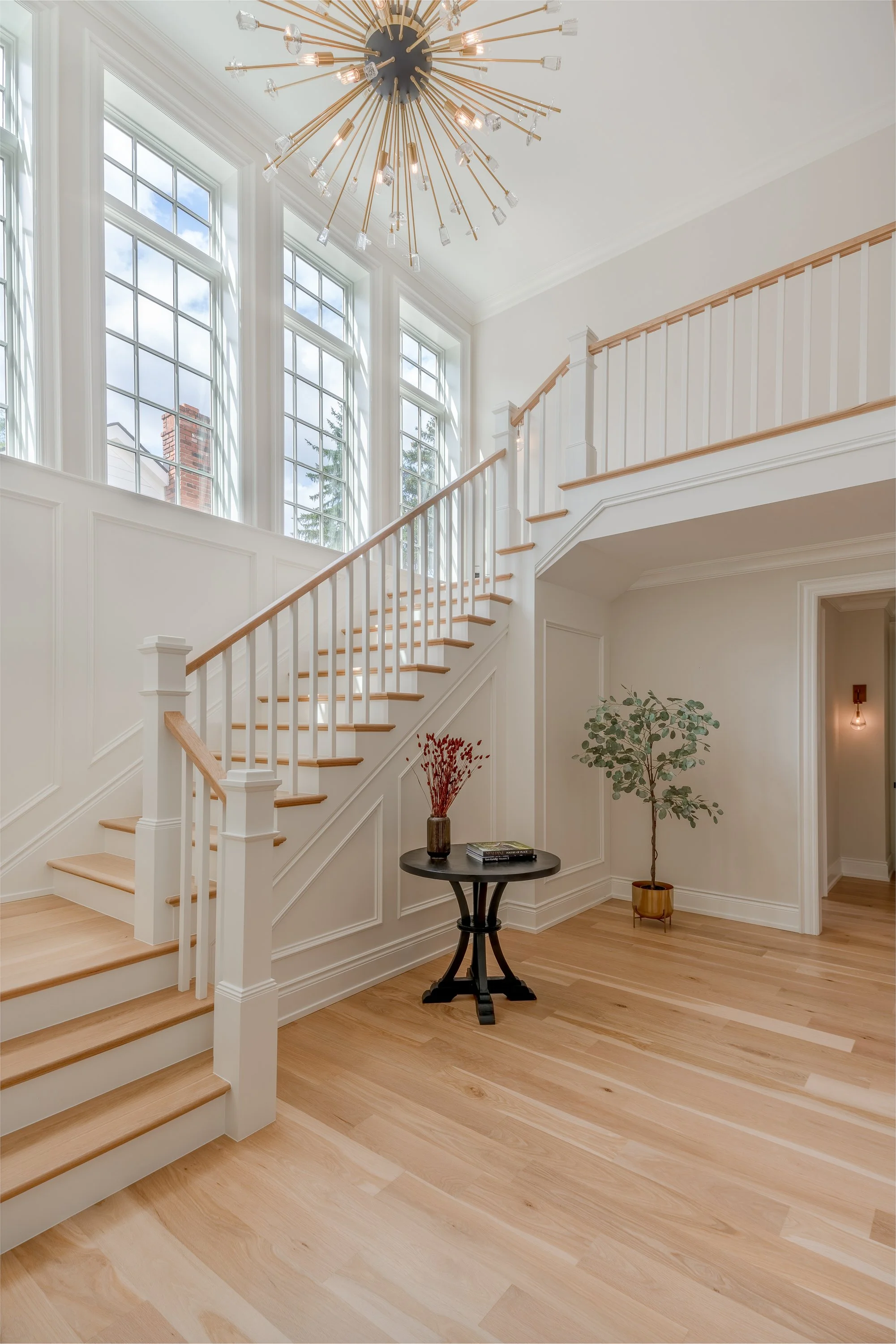 Bright entryway with a staircase, large windows, wood flooring, a black side table with a vase of dried flowers, and a potted plant.