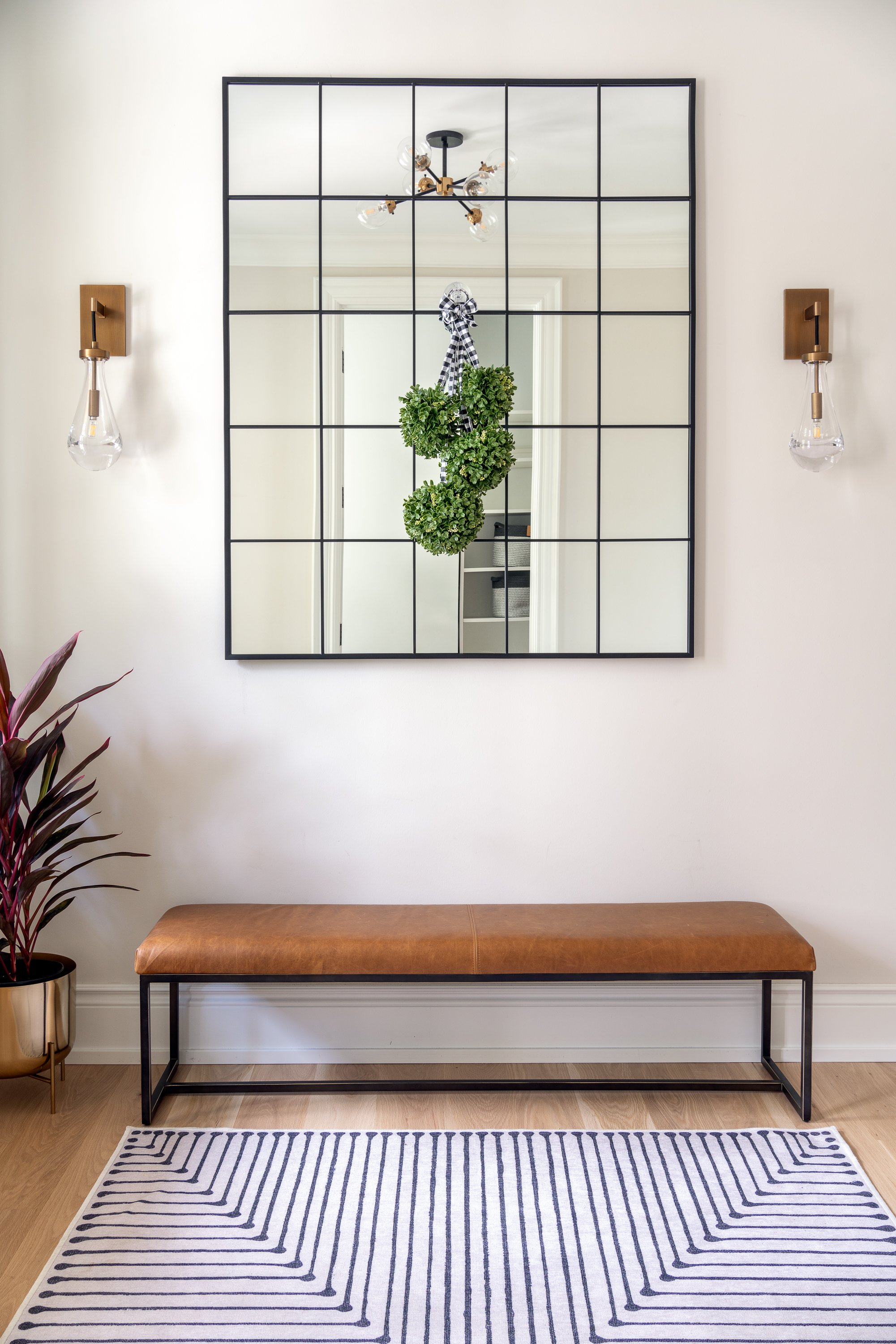 A mirror with a black grid frame reflecting a hanging plant with black and white checkered ribbon, two wall sconces with exposed bulbs, a brown bench with a black metal frame, a patterned rug, and a potted plant on a wooden floor.