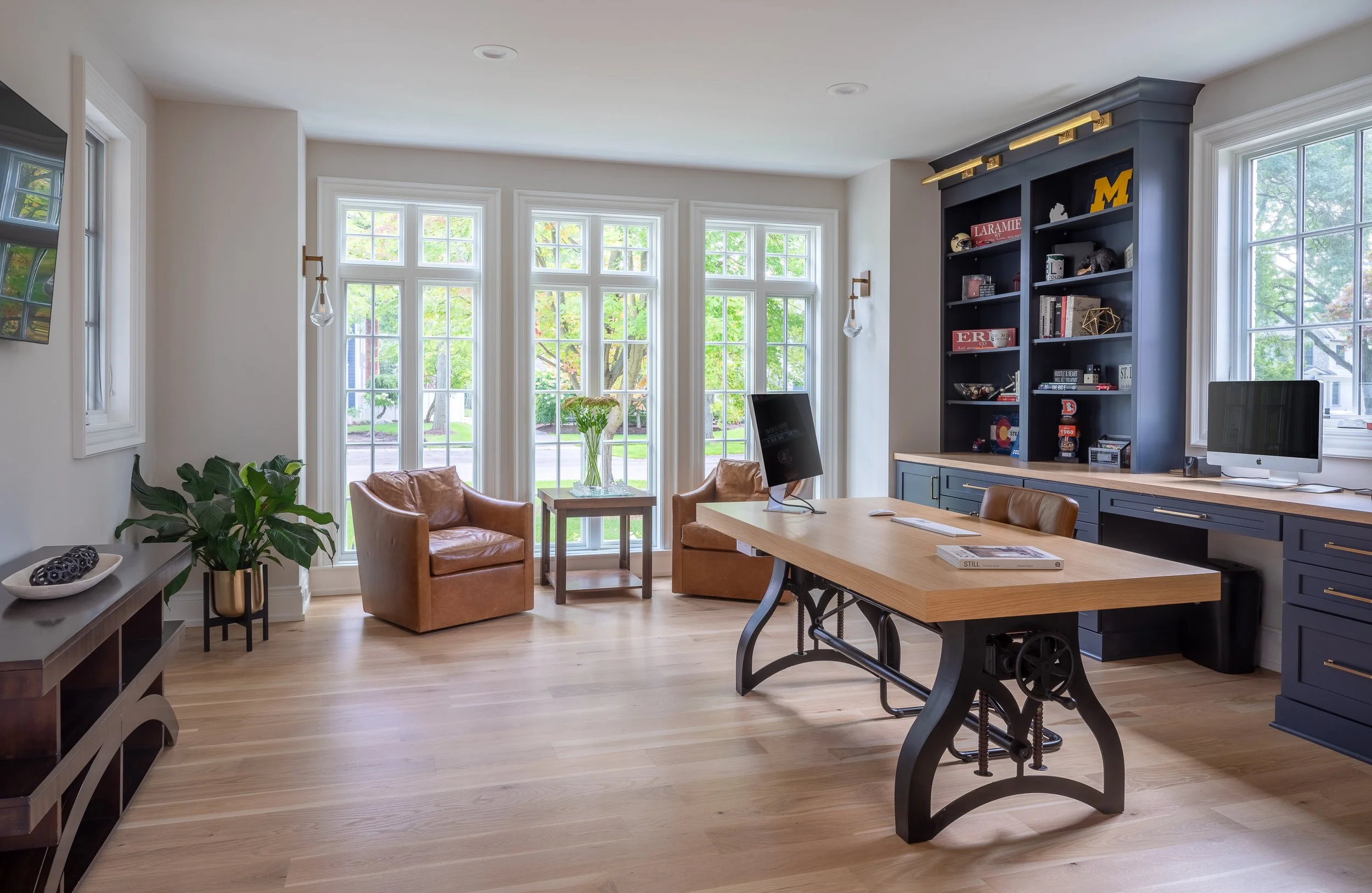 Bright home office with large windows, leather chairs, wooden desk, blue built-in shelves, and a computer