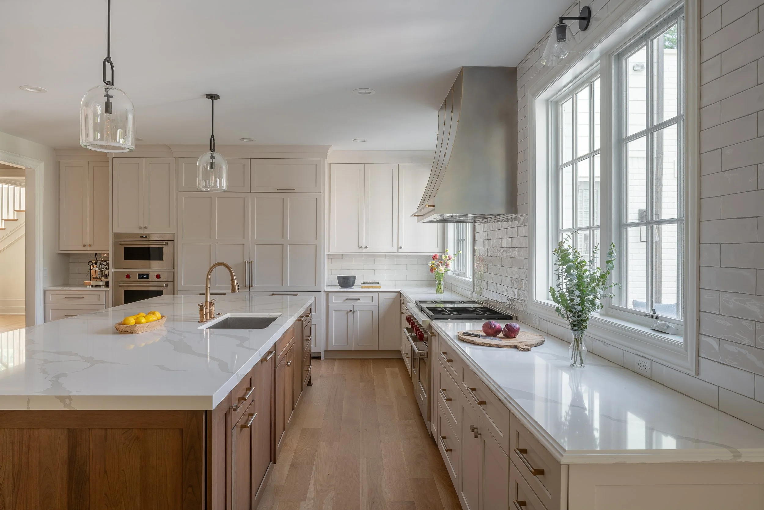 Modern kitchen with white cabinets, large island with a white countertop, stainless steel appliances, and large windows with a vase of greenery and a cutting board with red onions on the counter.