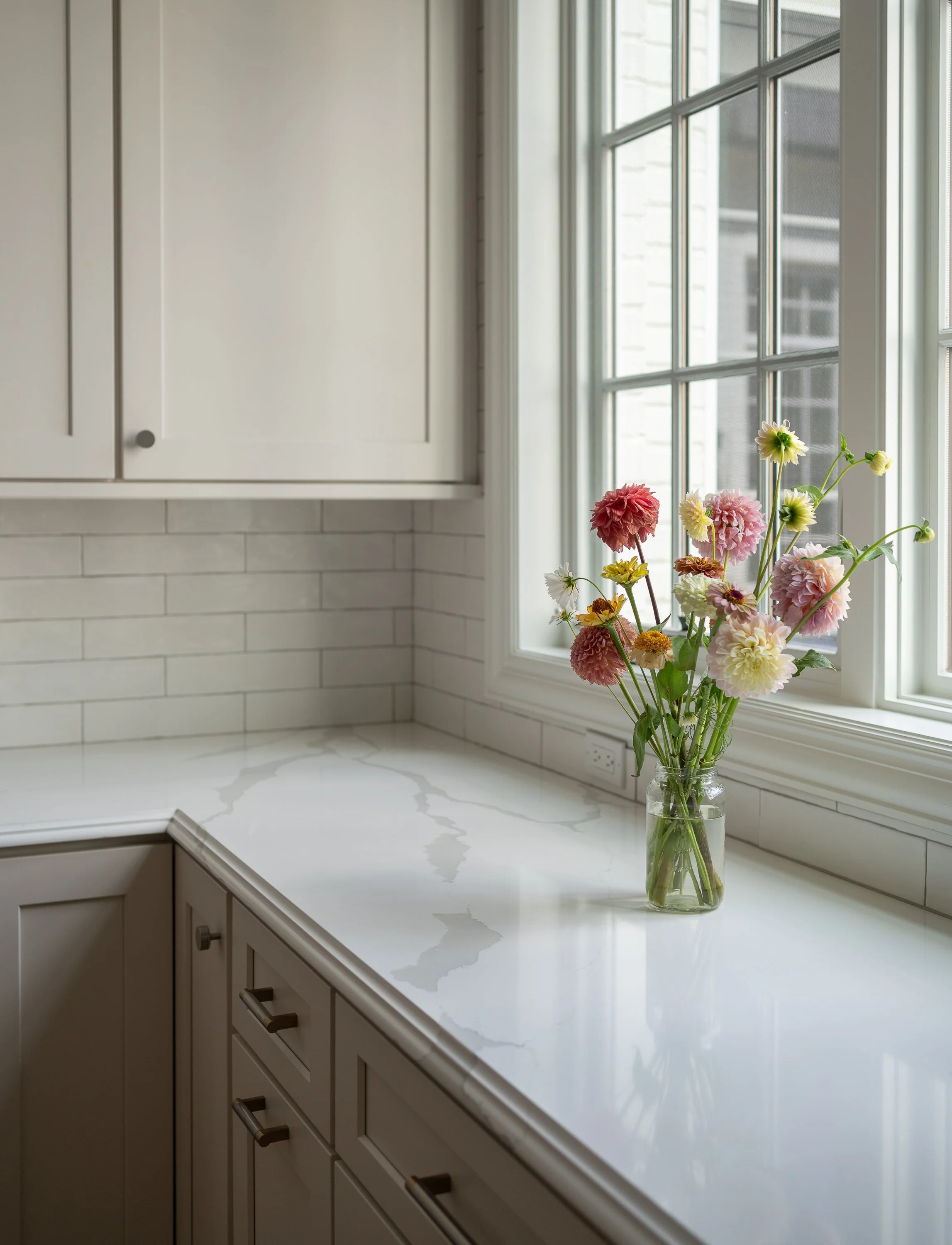 A white kitchen countertop with a marble pattern beside a window with white framing, holding a glass jar with pink, red, yellow, and white flowers.