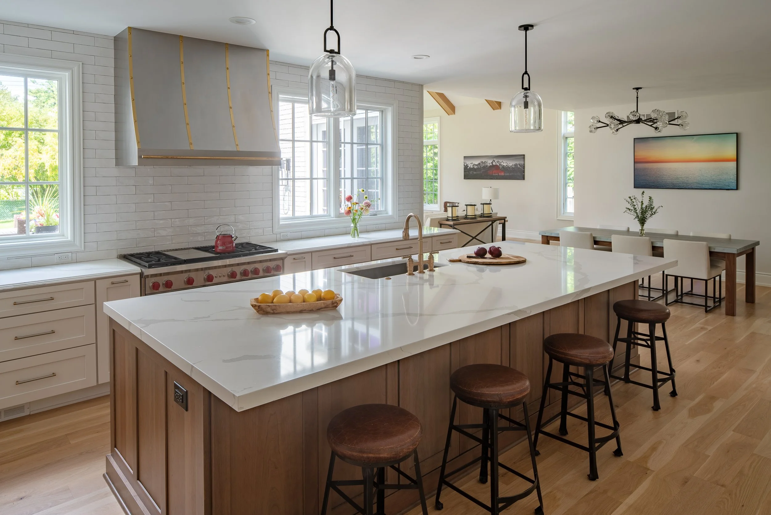 Modern kitchen featuring a large island with a white marble countertop and wooden base, four bar stools with dark metal frames and brown leather seats, a stainless steel gas stove with a red kettle, multiple windows letting in natural light, white ca