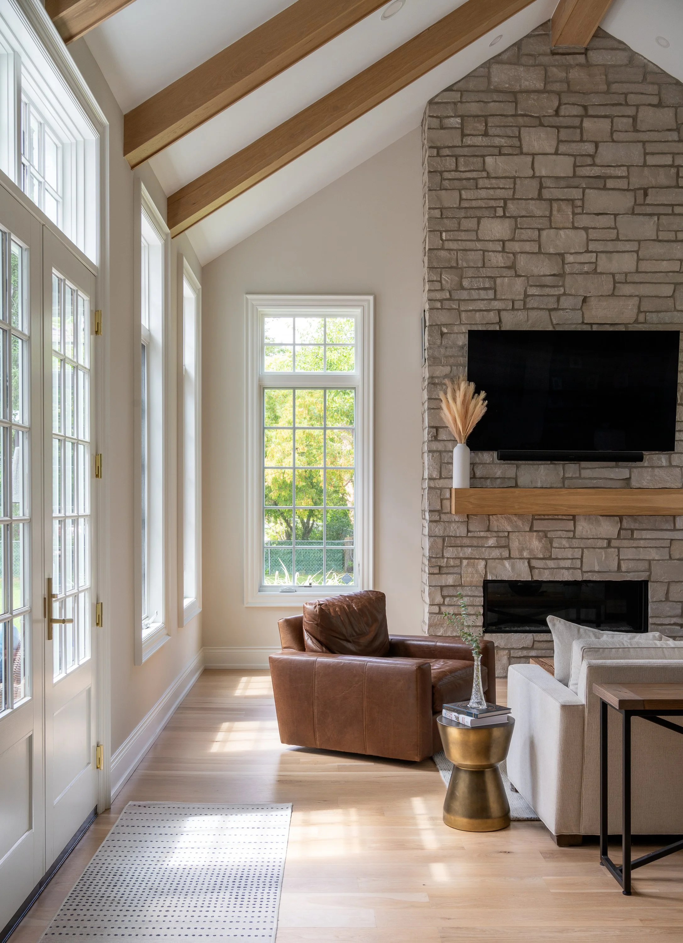 Living room with large windows, a stone fireplace with a mounted TV, a brown leather armchair, a beige sofa, a gold side table with books and a decorative plant, light wood flooring, and a white and black striped rug near a glass door.