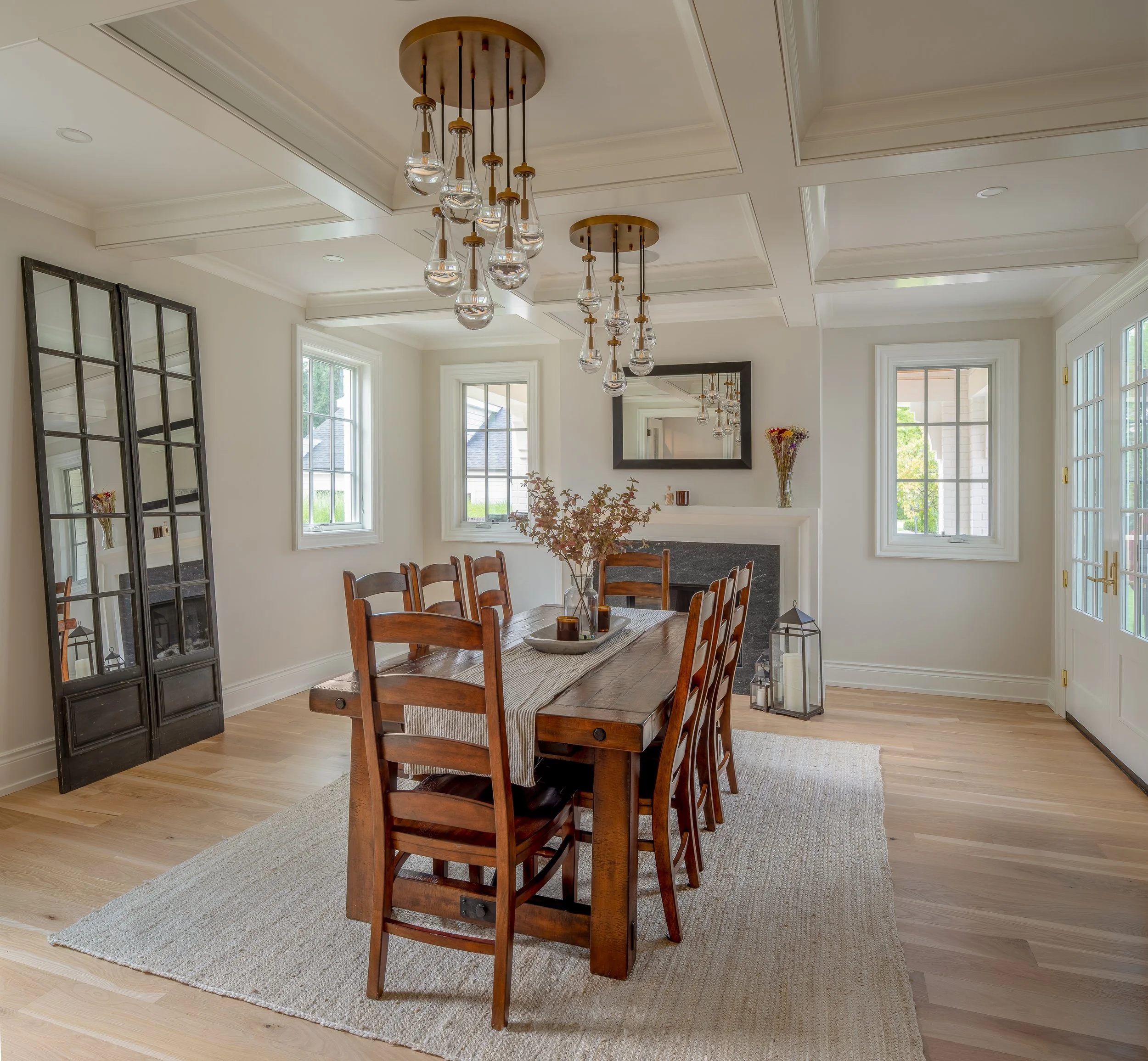 Dining room with wooden table and chairs, chandelier, large windows, mirror, fireplace with decorative items, lanterns, and a beige rug.