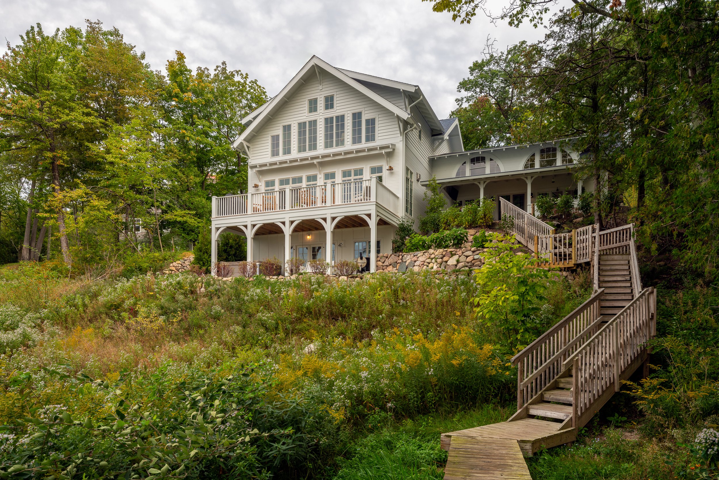 A large white house with multiple decks and stairs on a hillside, surrounded by trees and greenery.