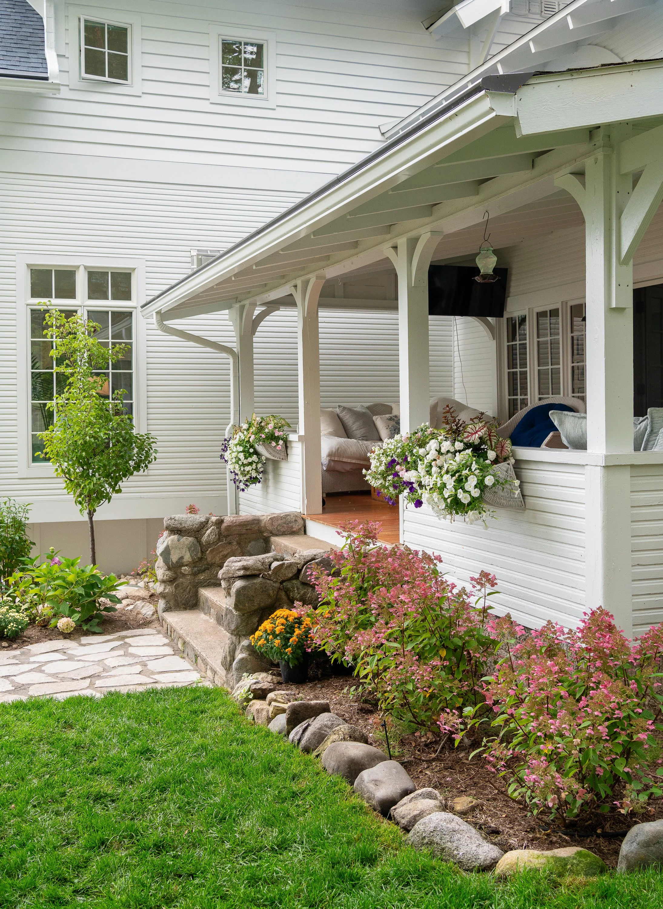 A front porch with white siding, a small set of stone steps, surrounded by a garden with green grass, pink, yellow, white, and purple flowers, and a potted plant. The porch has a hanging lantern, a mounted TV, and outdoor seating with cushions.
