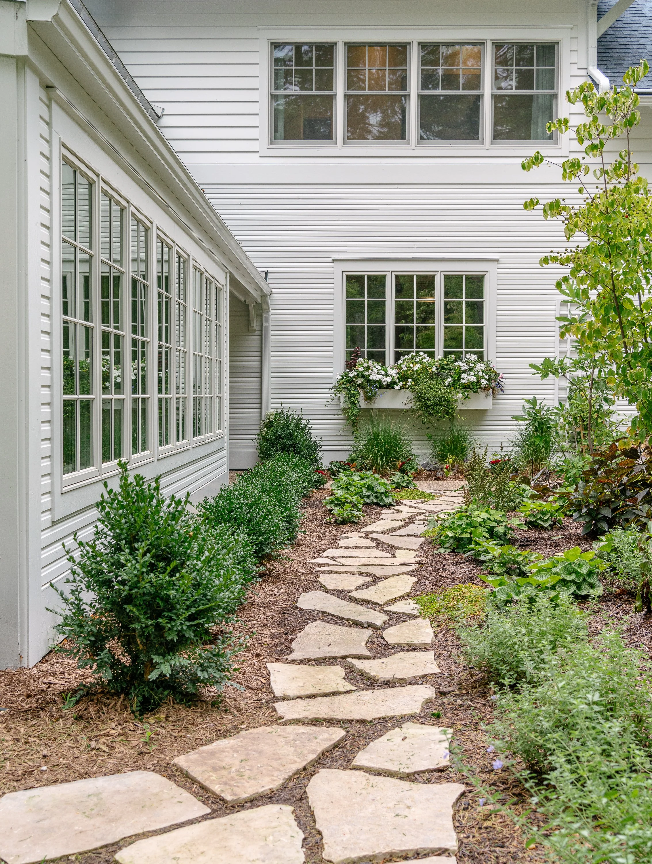Stone pathway leading to a white house with large windows and a garden with shrubs and flowers.