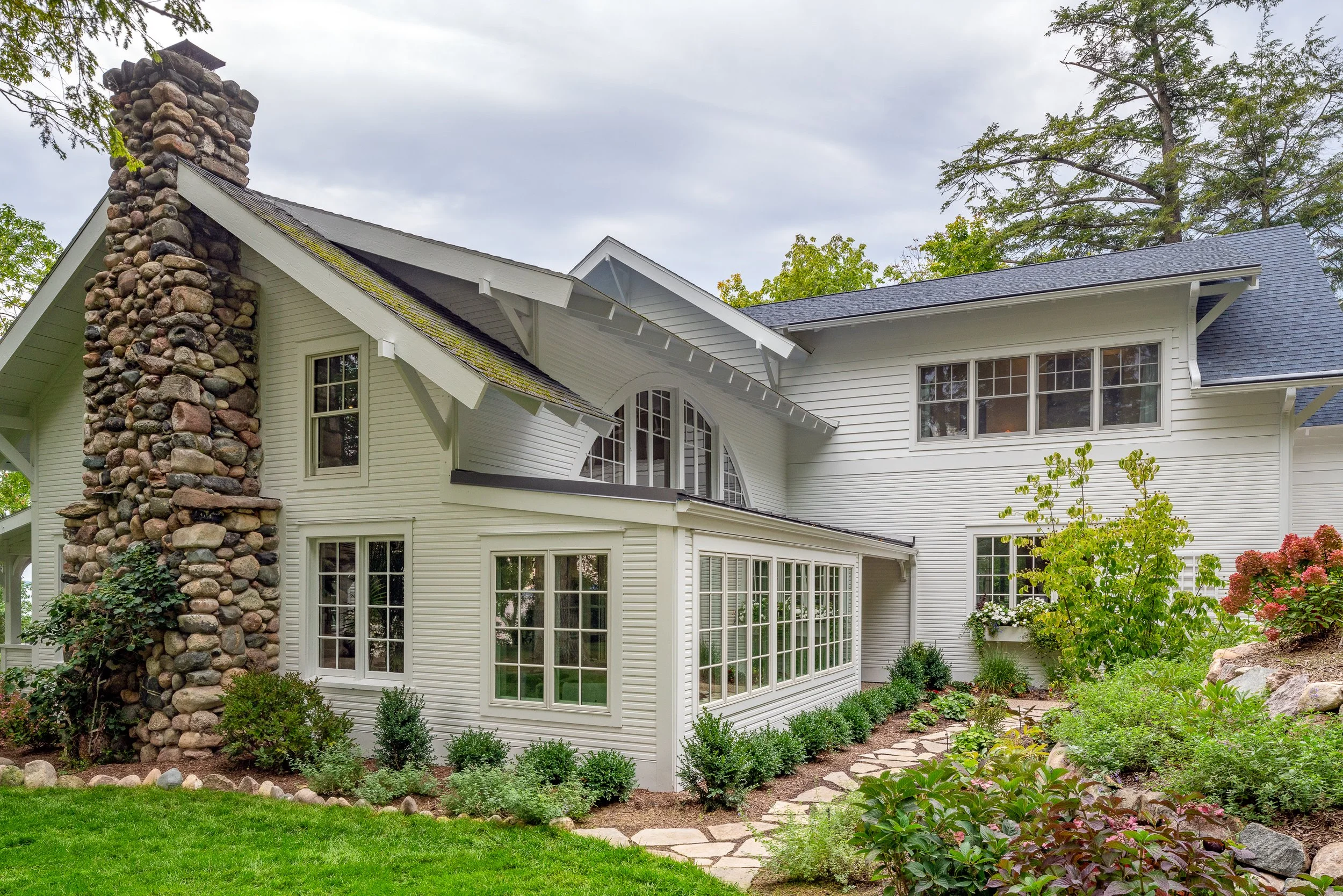 White house with large windows, stone chimney, and lush garden with plants and a stone pathway.