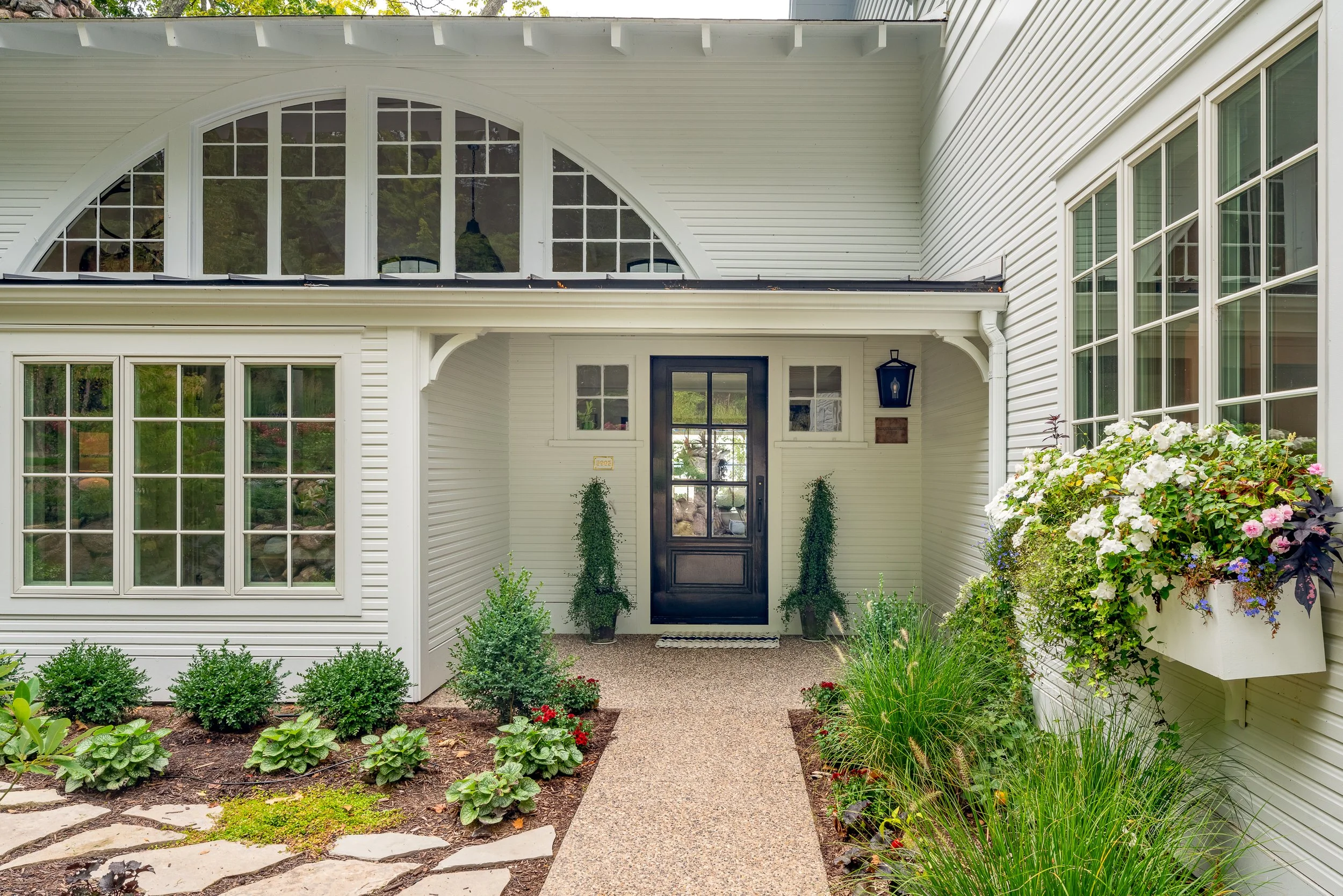 Front entrance of a house with a black door, surrounded by white siding, large windows, and landscaped garden with bushes, flowers, and potted plants.