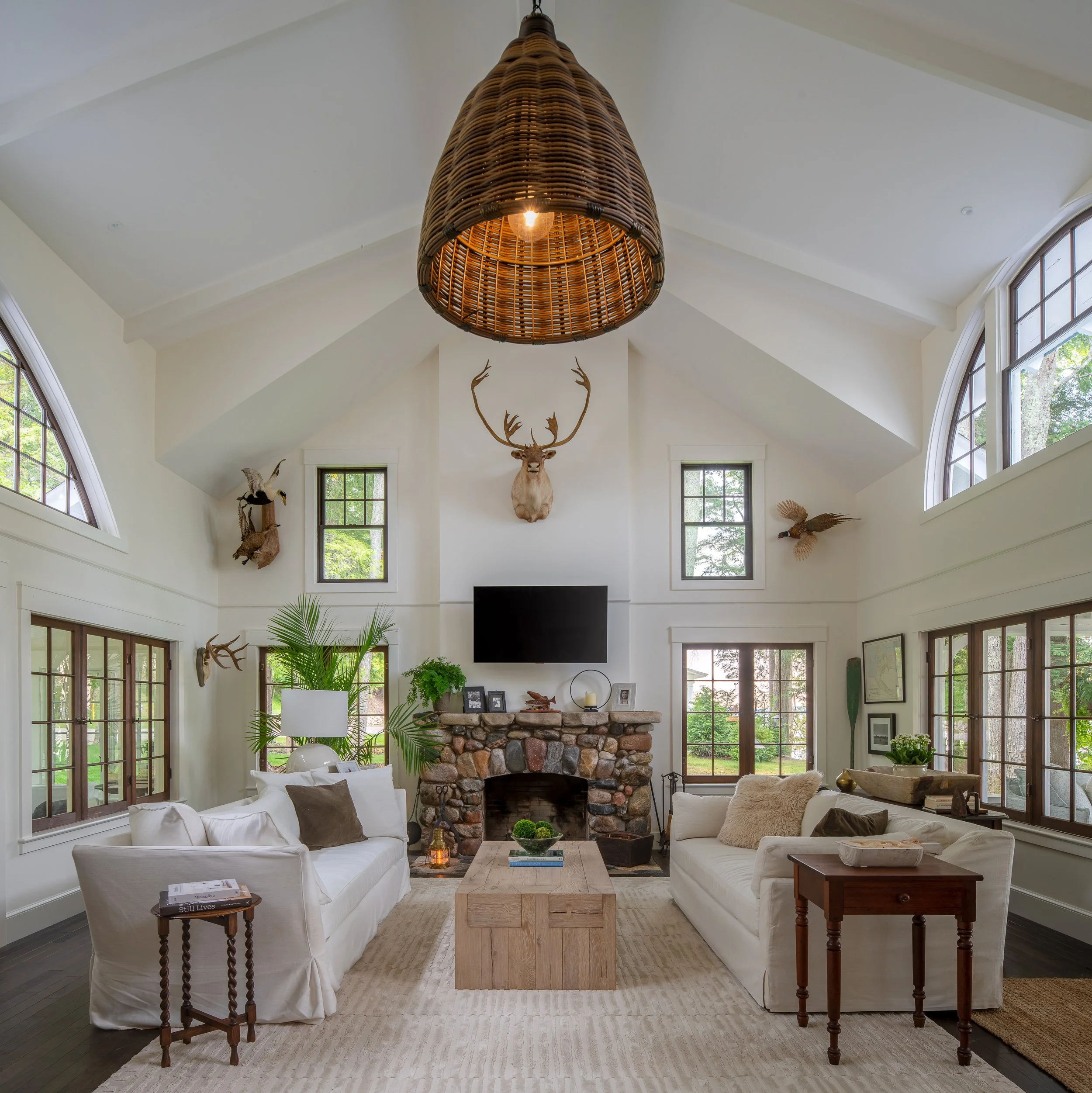 Living room with white sofas, a stone fireplace, large windows, mounted animal heads, and wooden furniture.