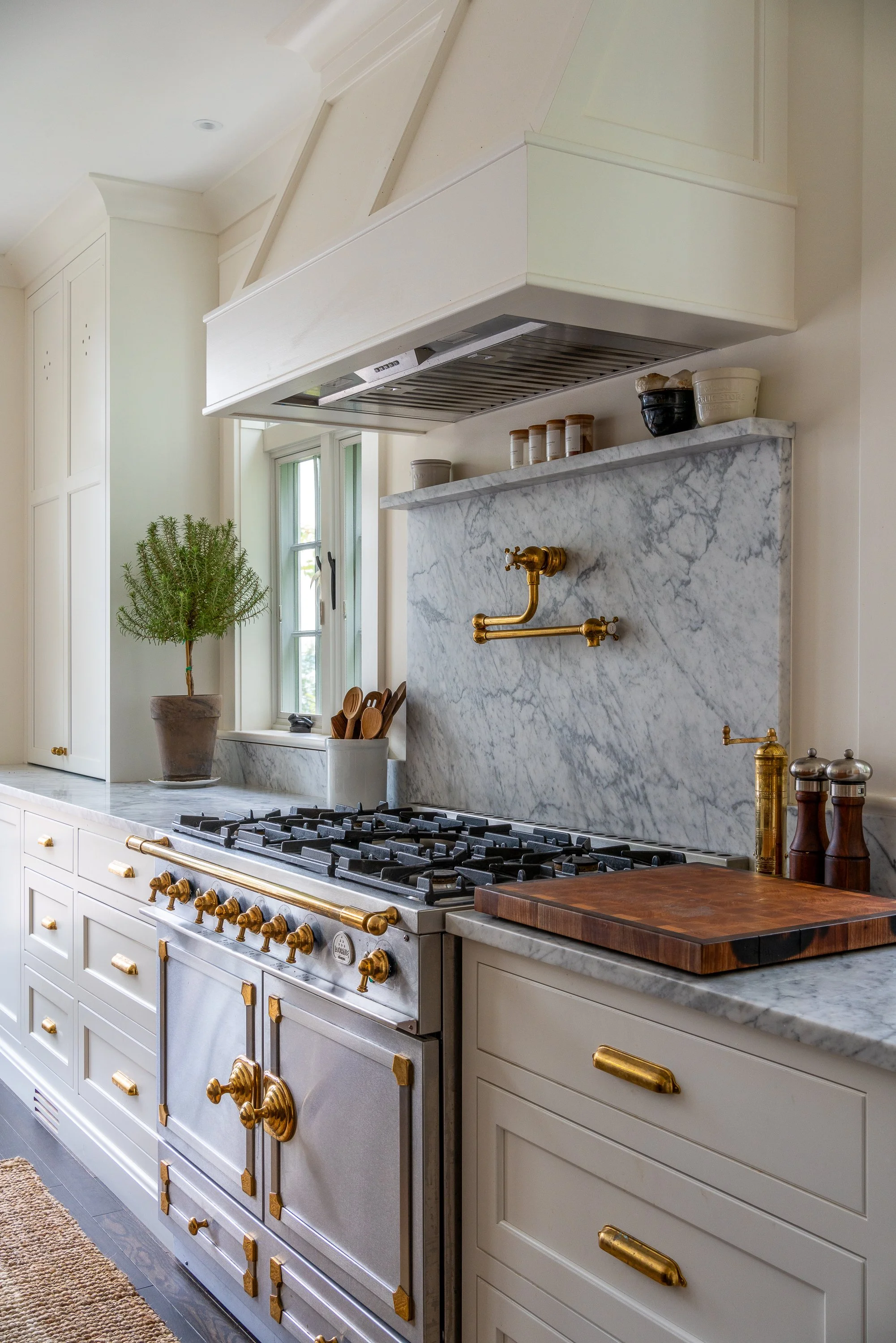 A kitchen with white cabinets, a marble backsplash, a gas stove with gold accents, a wooden cutting board, a potted plant, and a window with utensils nearby.