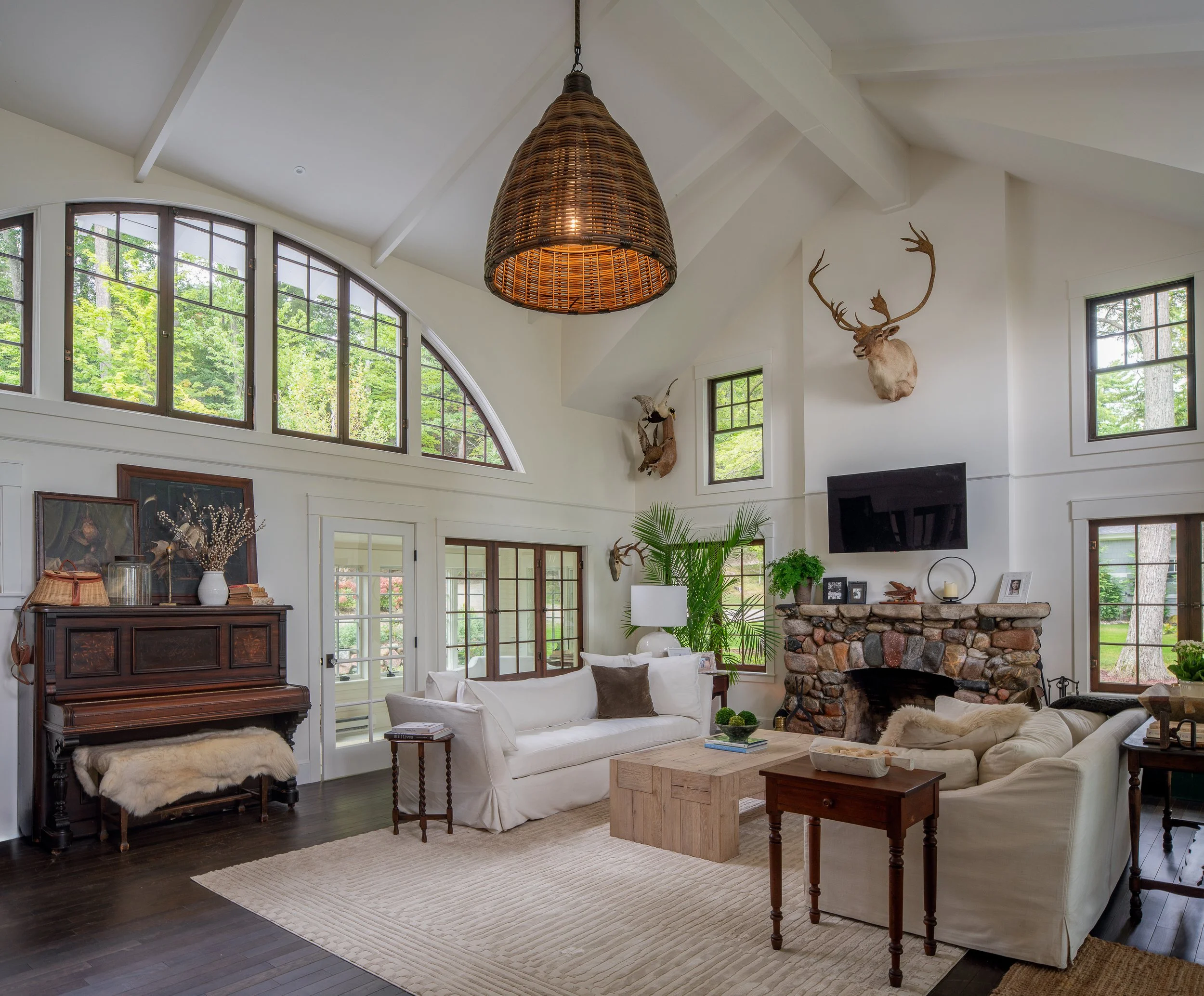 Living room with white sofas, a stone fireplace, large windows, mounted animal heads, a hanging rattan light fixture, and various decorative items.