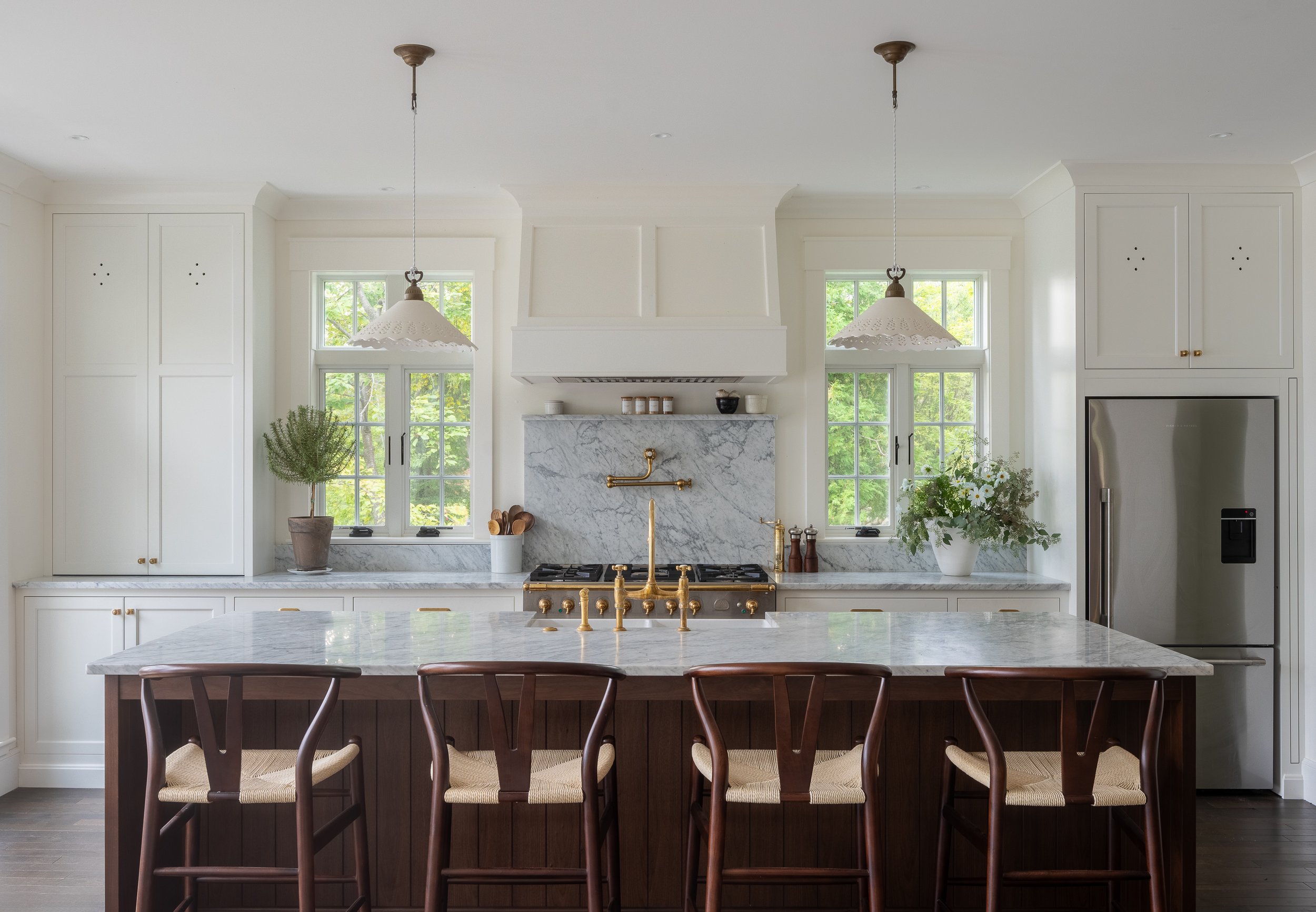 Modern kitchen with white cabinetry, marble countertops, a central island with dark wood base, and brass fixtures. There are two windows with greenery outside, hanging pendant lights, and a stainless steel refrigerator.