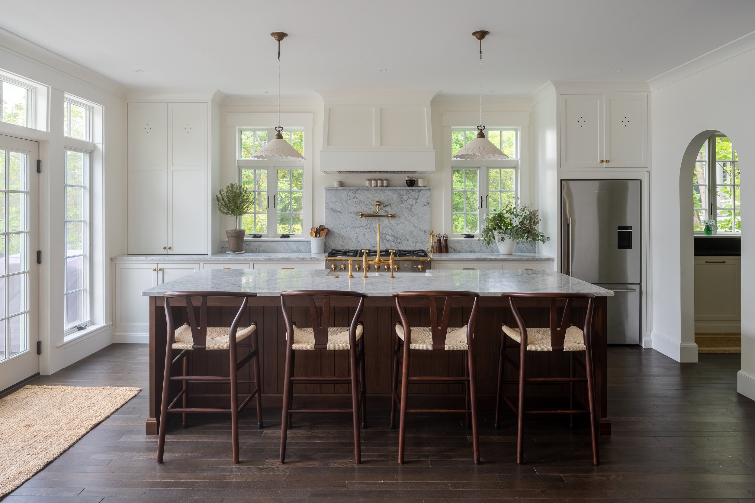 Modern kitchen with white cabinets, marble countertops, a kitchen island with a marble top and wooden base, dark wood floors, and large windows with greenery outside.