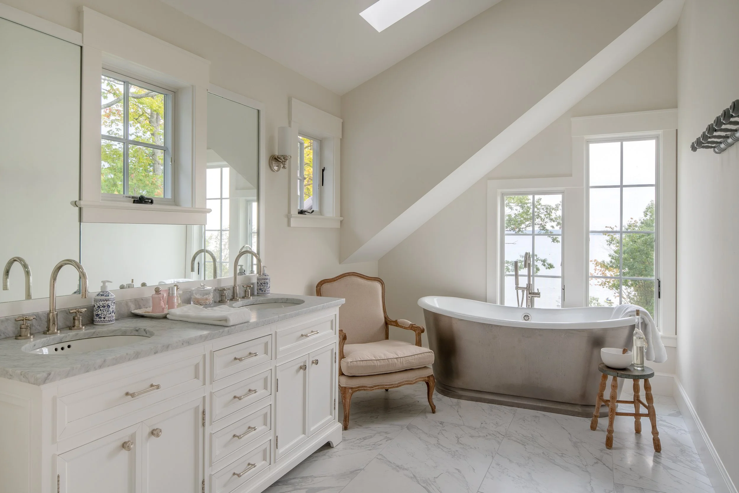 Bright bathroom with a double sink vanity, a bathtub near large windows, and a vintage chair.