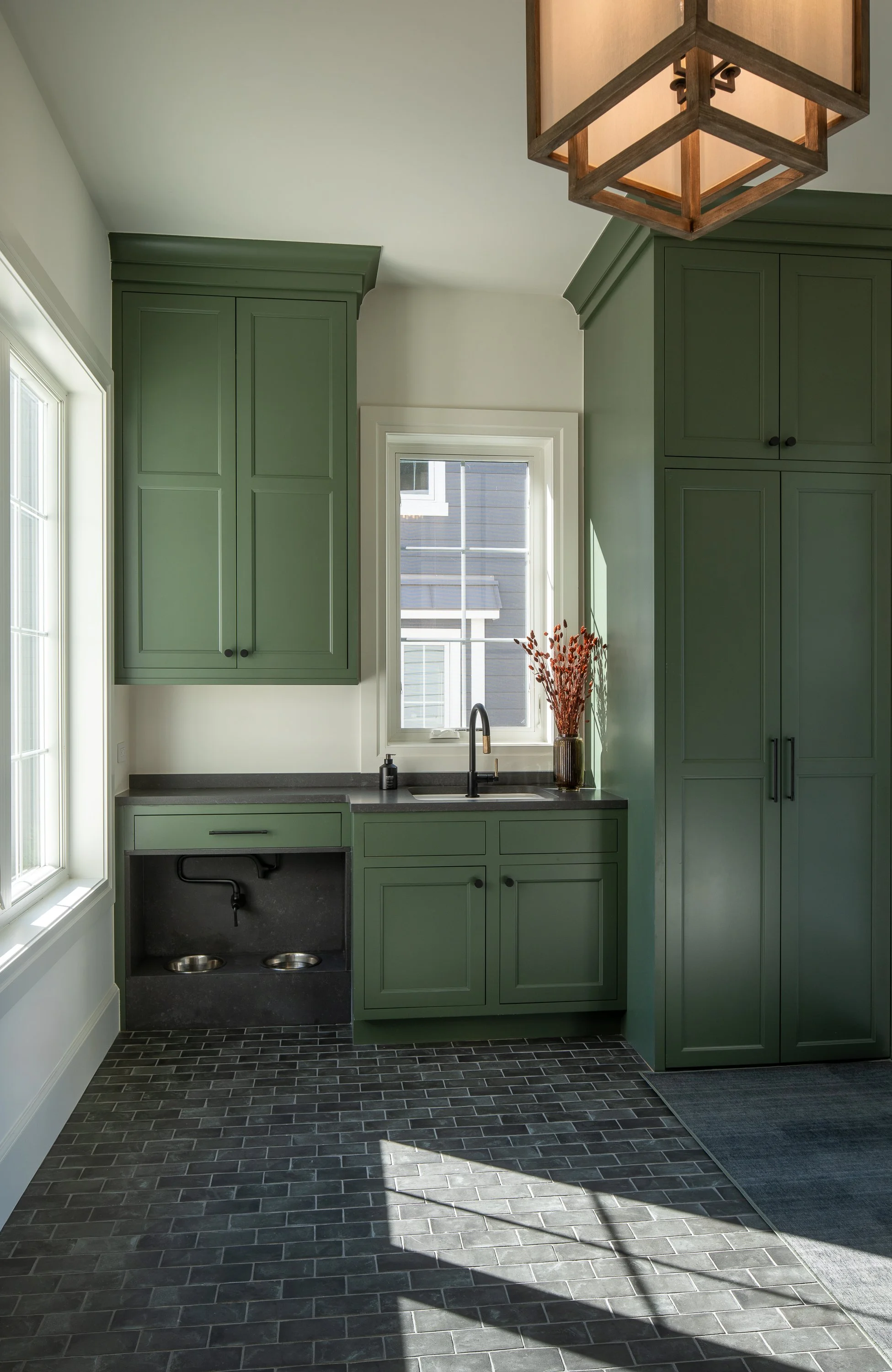 Interior of a kitchen corner with green cabinets, a black sink, gray countertop, a window with exterior view, and sunlight casting shadows on a dark brick floor.