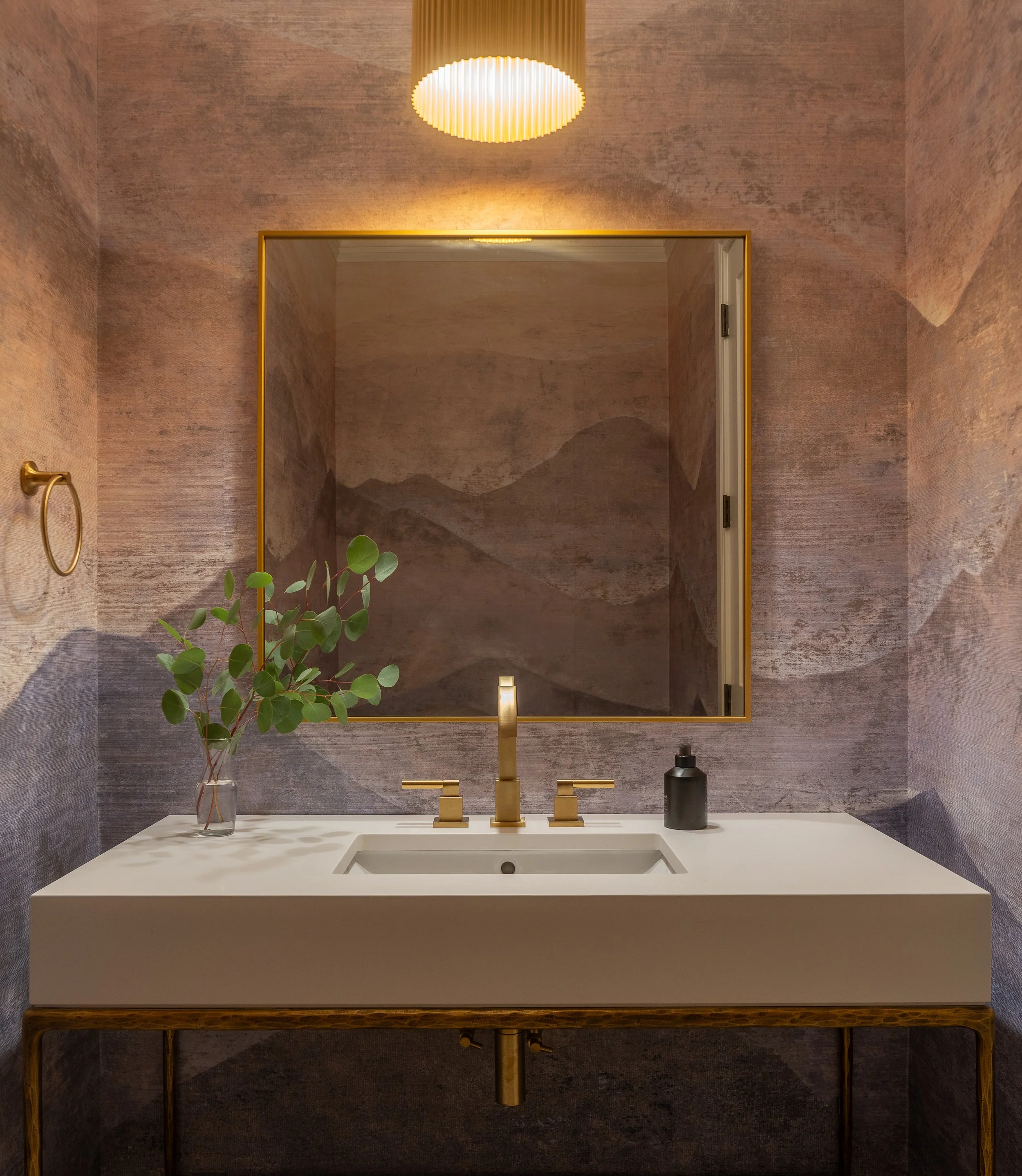 Bathroom sink with gold fixtures, a mirror, a black soap dispenser, and a vase with green leaves, against a textured wall with mountain-like art.