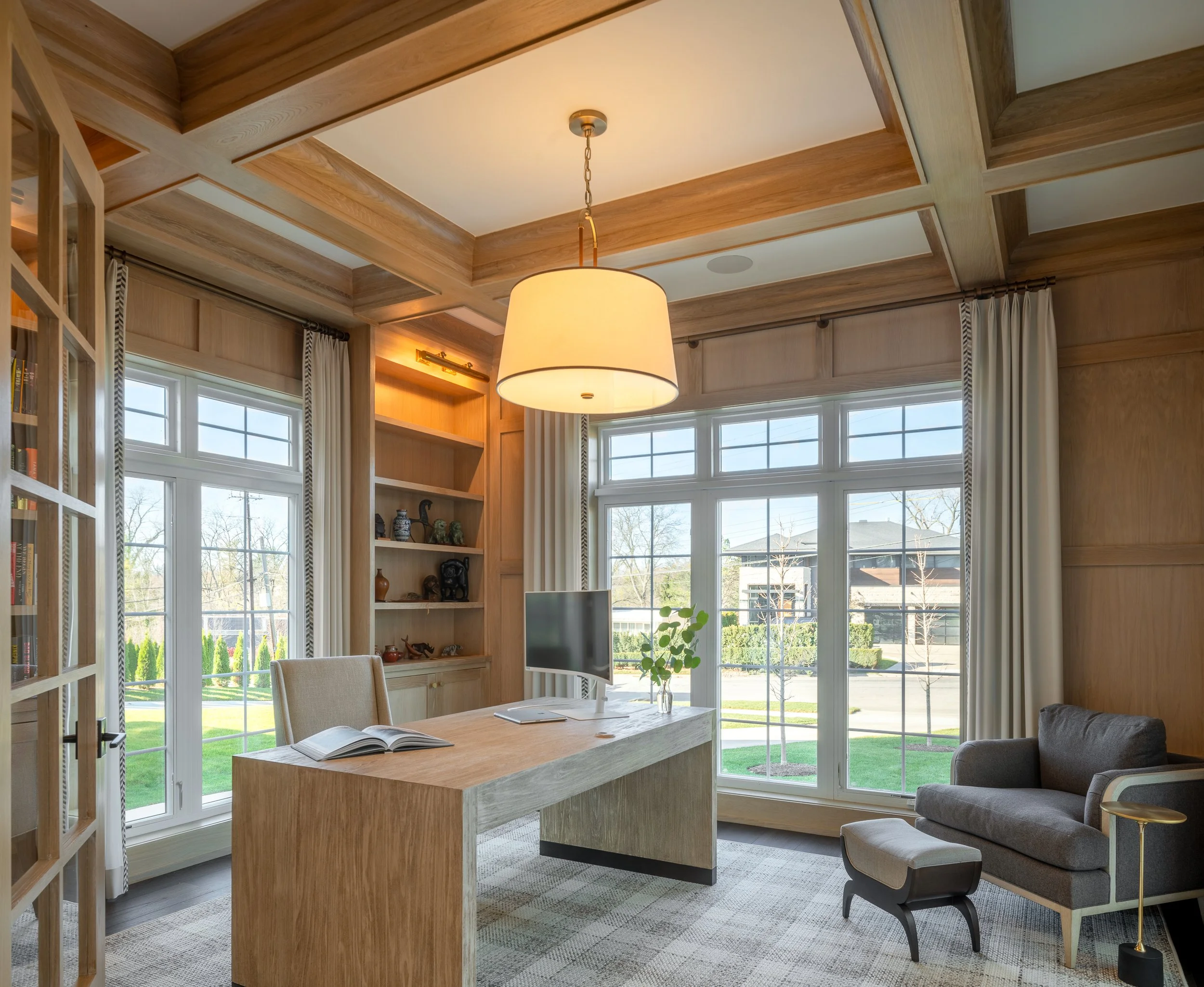 Bright home office with large windows, wooden ceiling, beige desk, gray sofa, bookshelf, and a large hanging light.