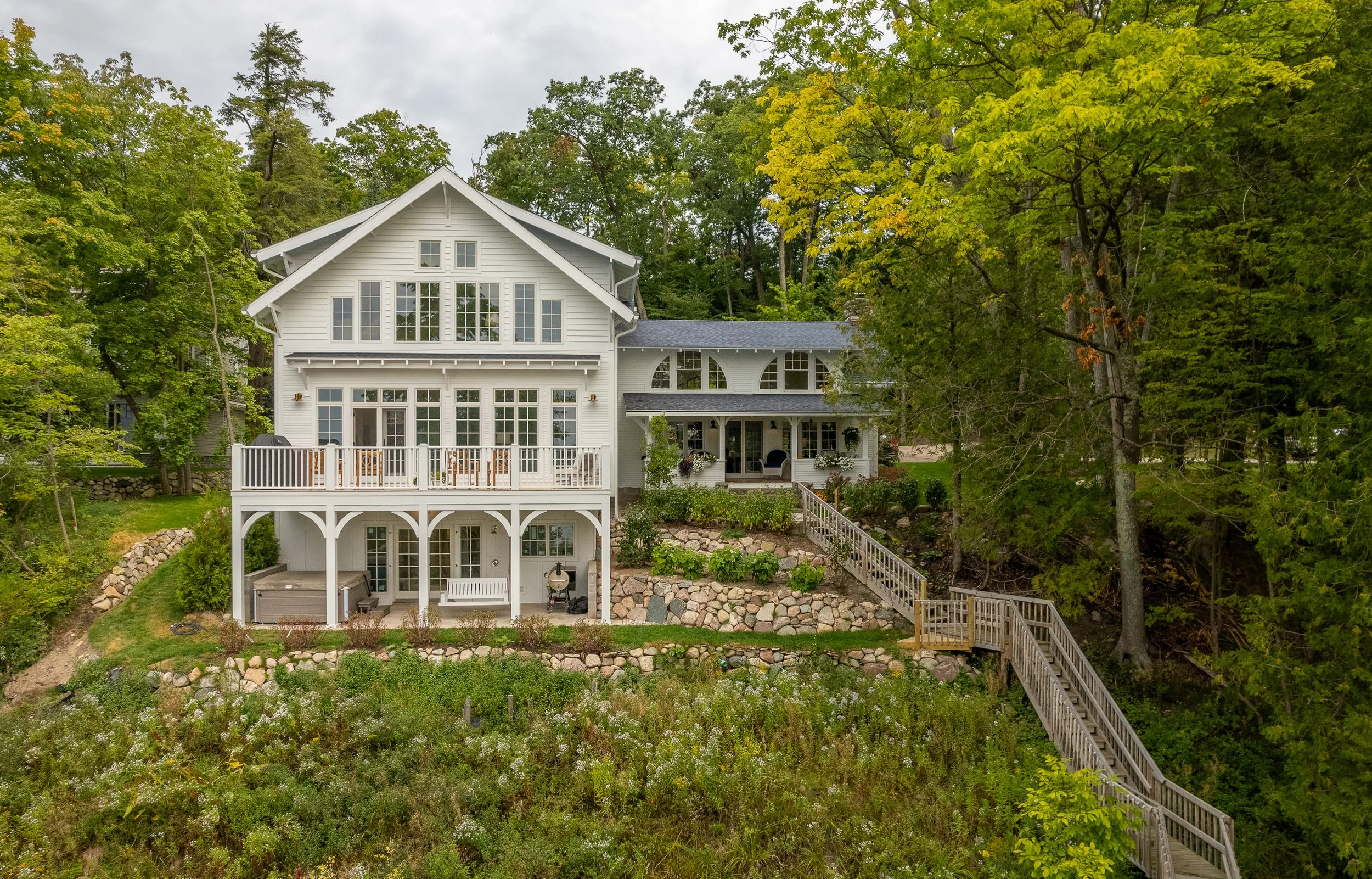 Large white house on a hill with wooden stairs leading up to it, surrounded by green trees and natural landscaping.