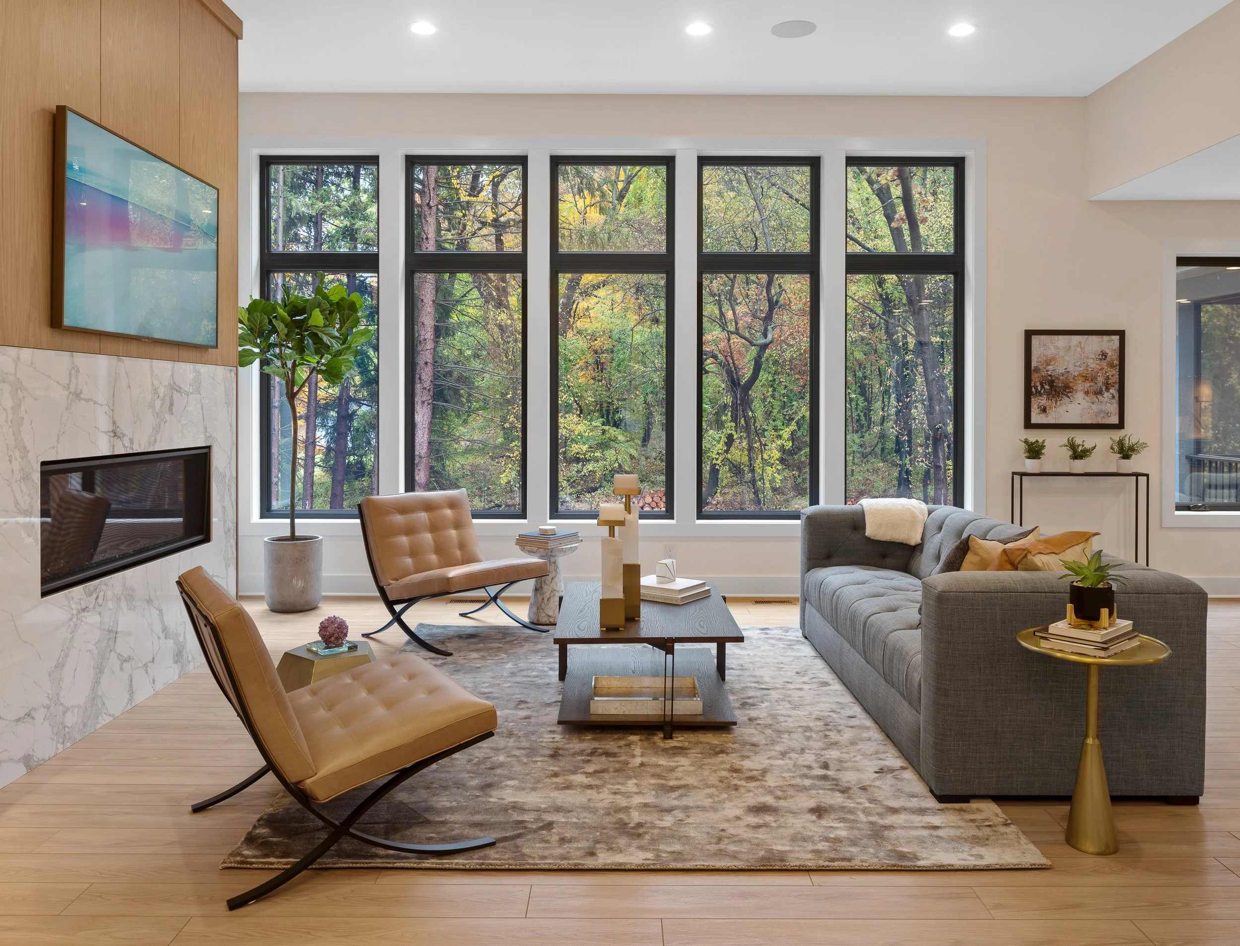 Modern living room with large windows overlooking a wooded outdoor scene. Features a gray sofa, tan chairs, a marble fireplace, a flat-screen TV, and gold side tables, with a rug and decorative plants.