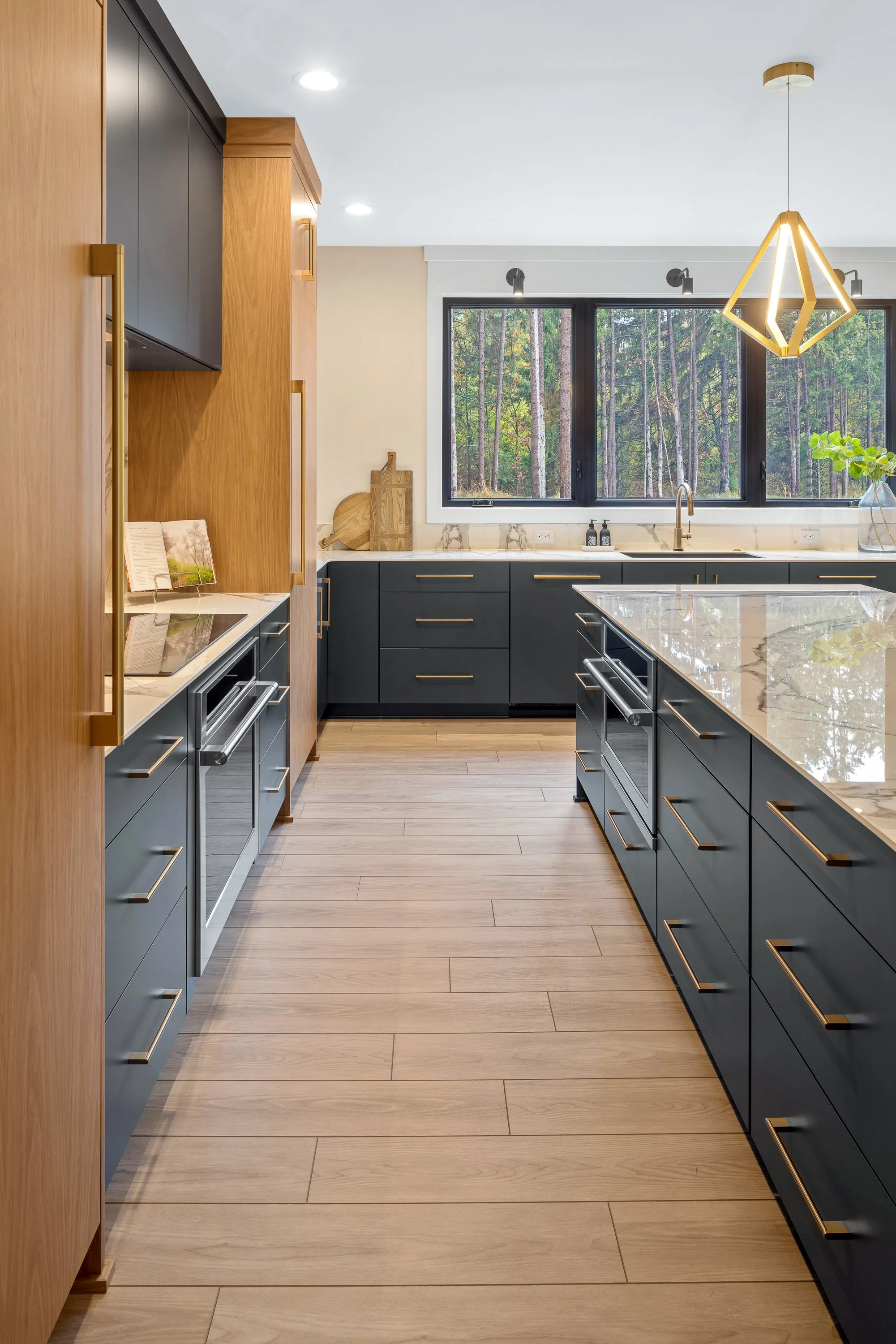 Modern kitchen with black and wood cabinets, marble countertops, light wood flooring, and large window overlooking a forest.
