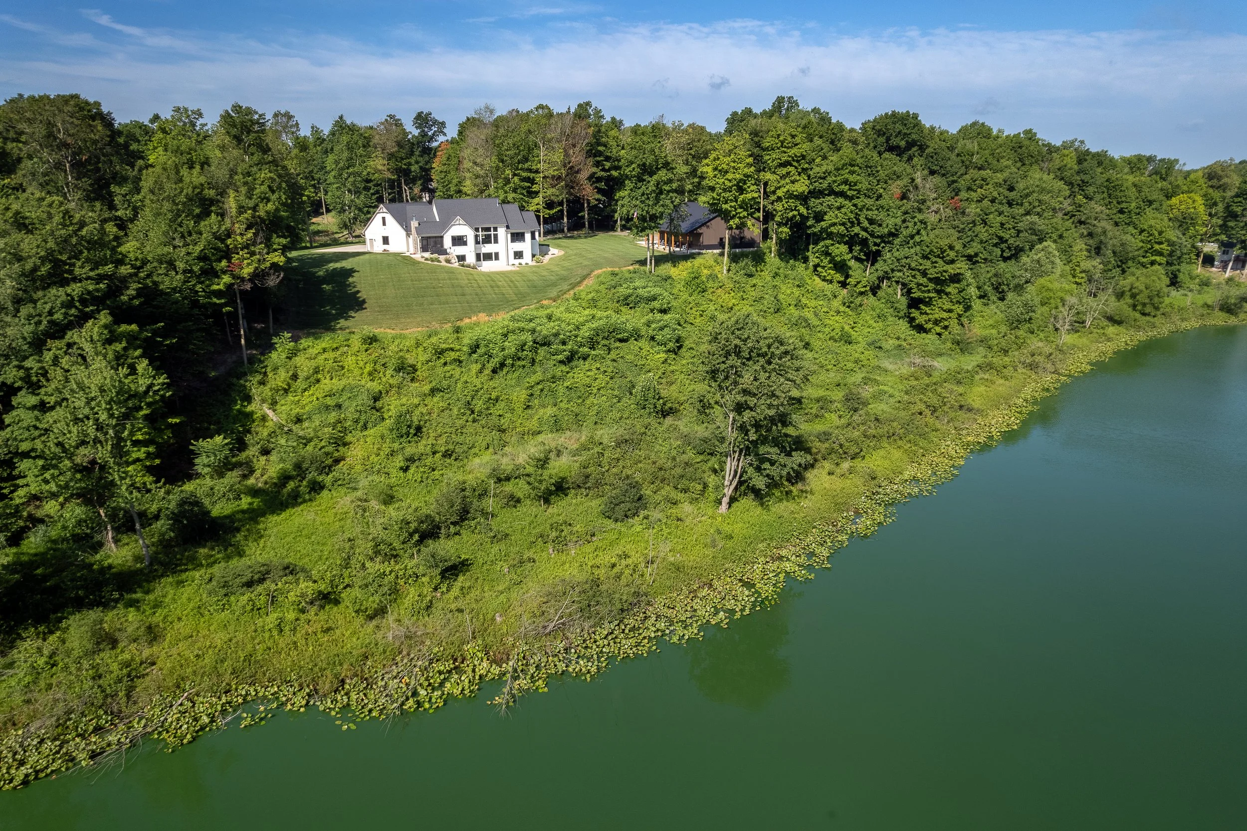 A large house with a gray roof situated on a green hill overlooking a body of water surrounded by lush trees.