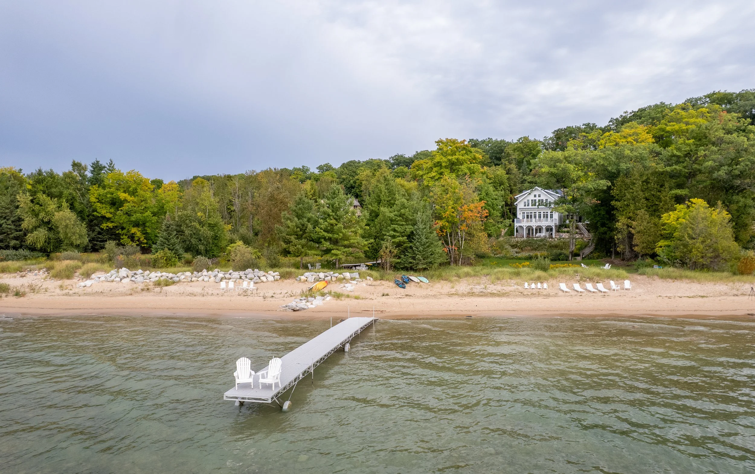 A white dock with chairs extends into a lake, with a sandy beach and green trees in the background, and a large house on a hill.