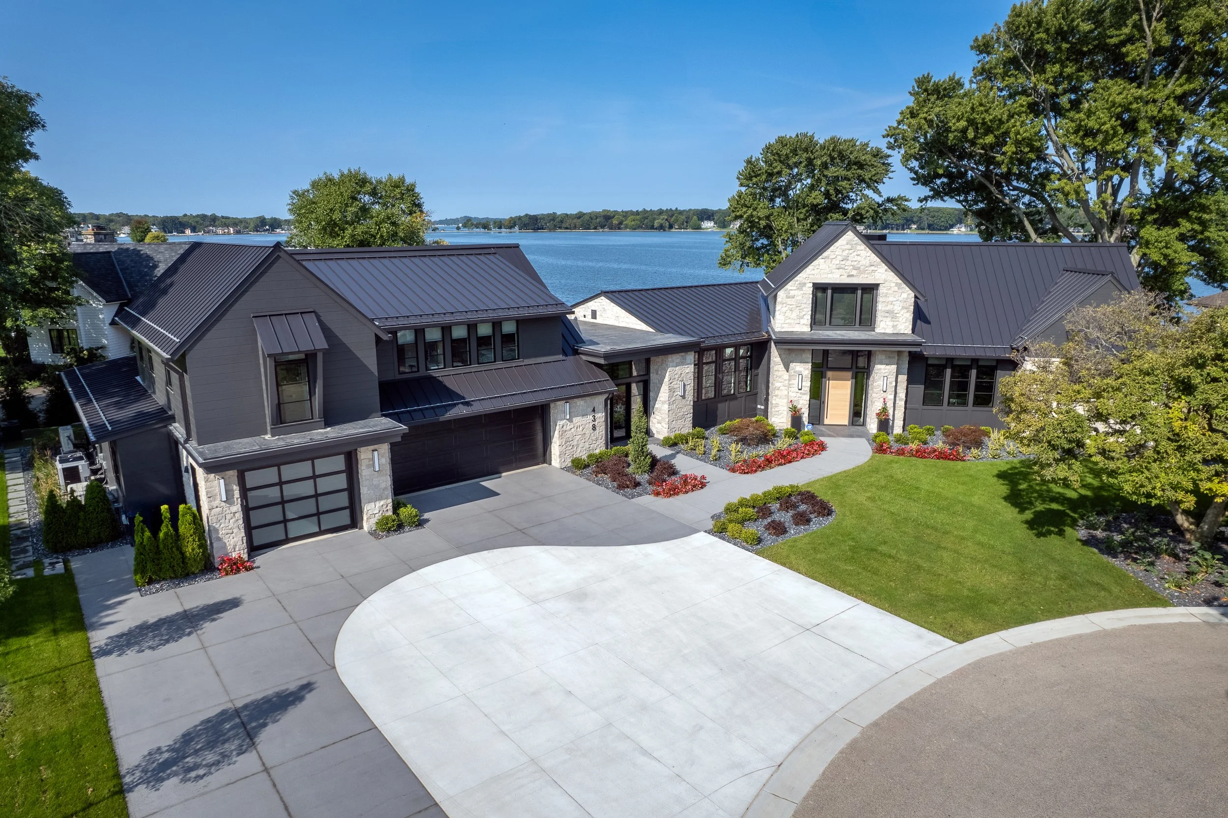 Modern house with gray and stone exterior, black roof, large driveway, lush front yard, and trees, with a lakeside view in background on a sunny day.
