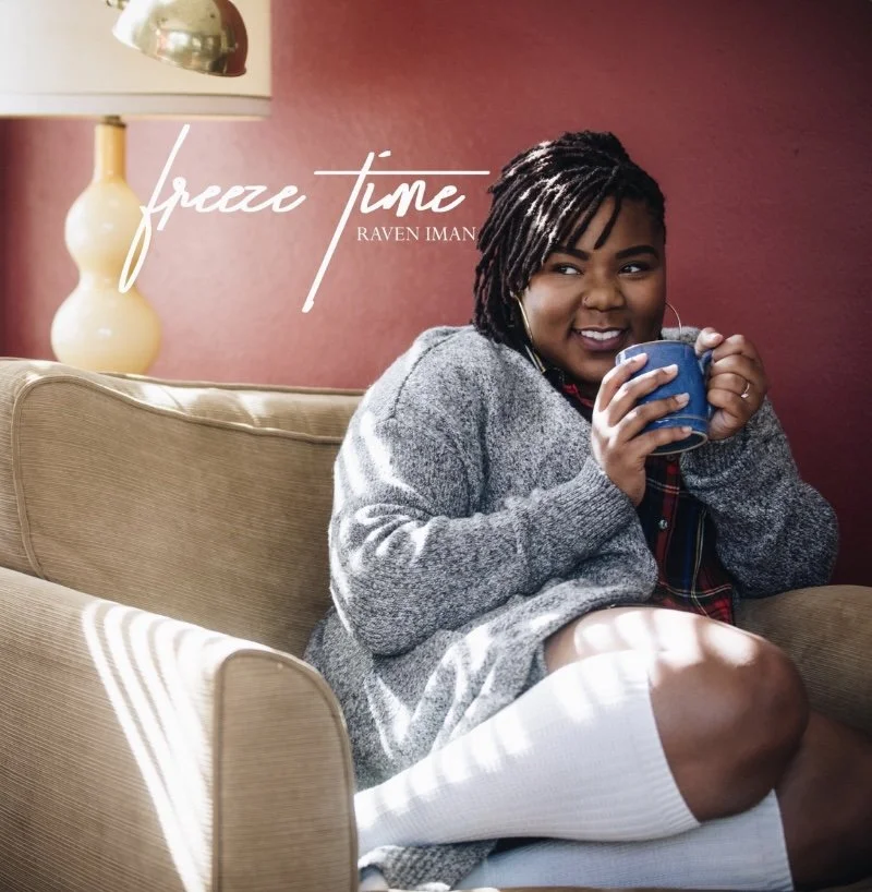 A woman with dreadlocks sitting on a beige couch, holding a blue mug, smiling, with a maroon wall and a yellow lamp in the background. Text on the image reads "Freeze Time" by Raven Iman.