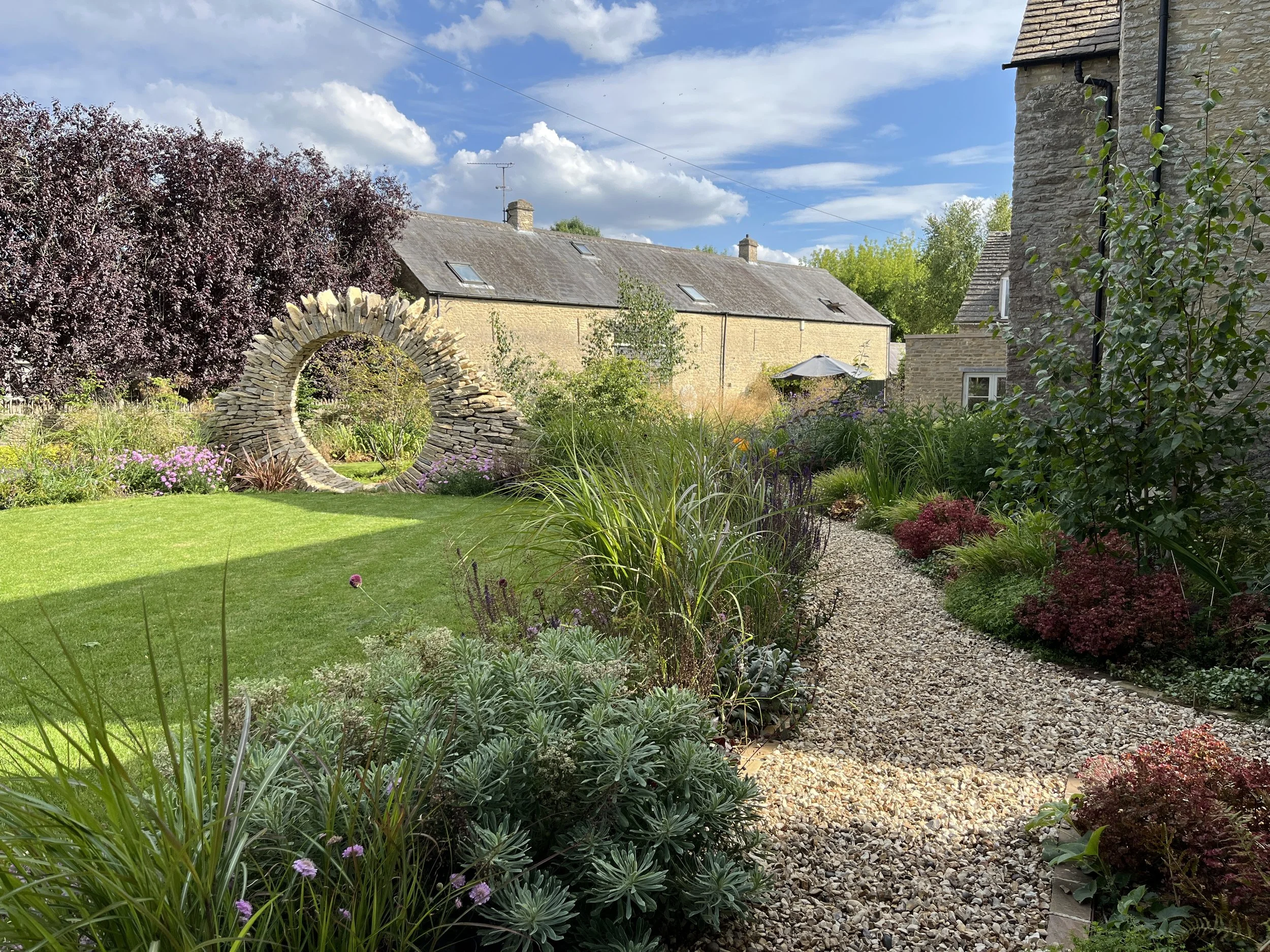 A lush garden with a gravel pathway, colorful flowering plants, trees, and a stone arch, with traditional brick houses and a blue sky with clouds in the background.