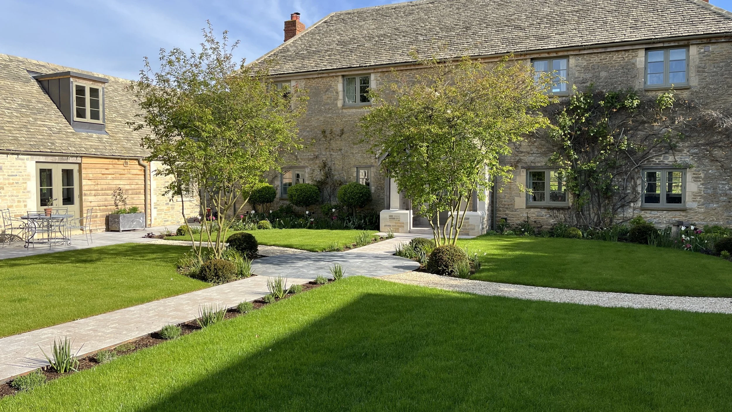 Well-maintained garden with lush green lawn, stone pathway, small trees, and a stone house with multiple windows and climbing plants, under a clear blue sky.