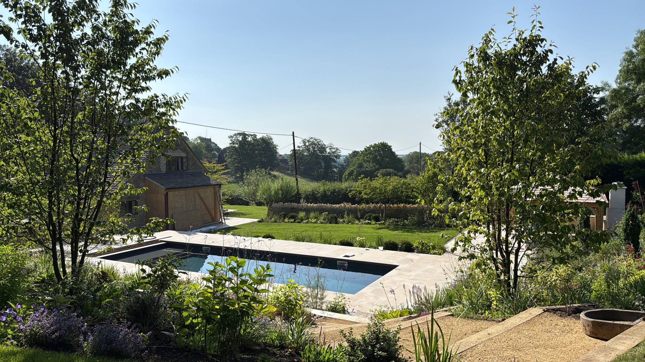 A garden with a swimming pool, surrounded by lush green grass, trees, and shrubs, with rolling hills and clear blue sky in the background.