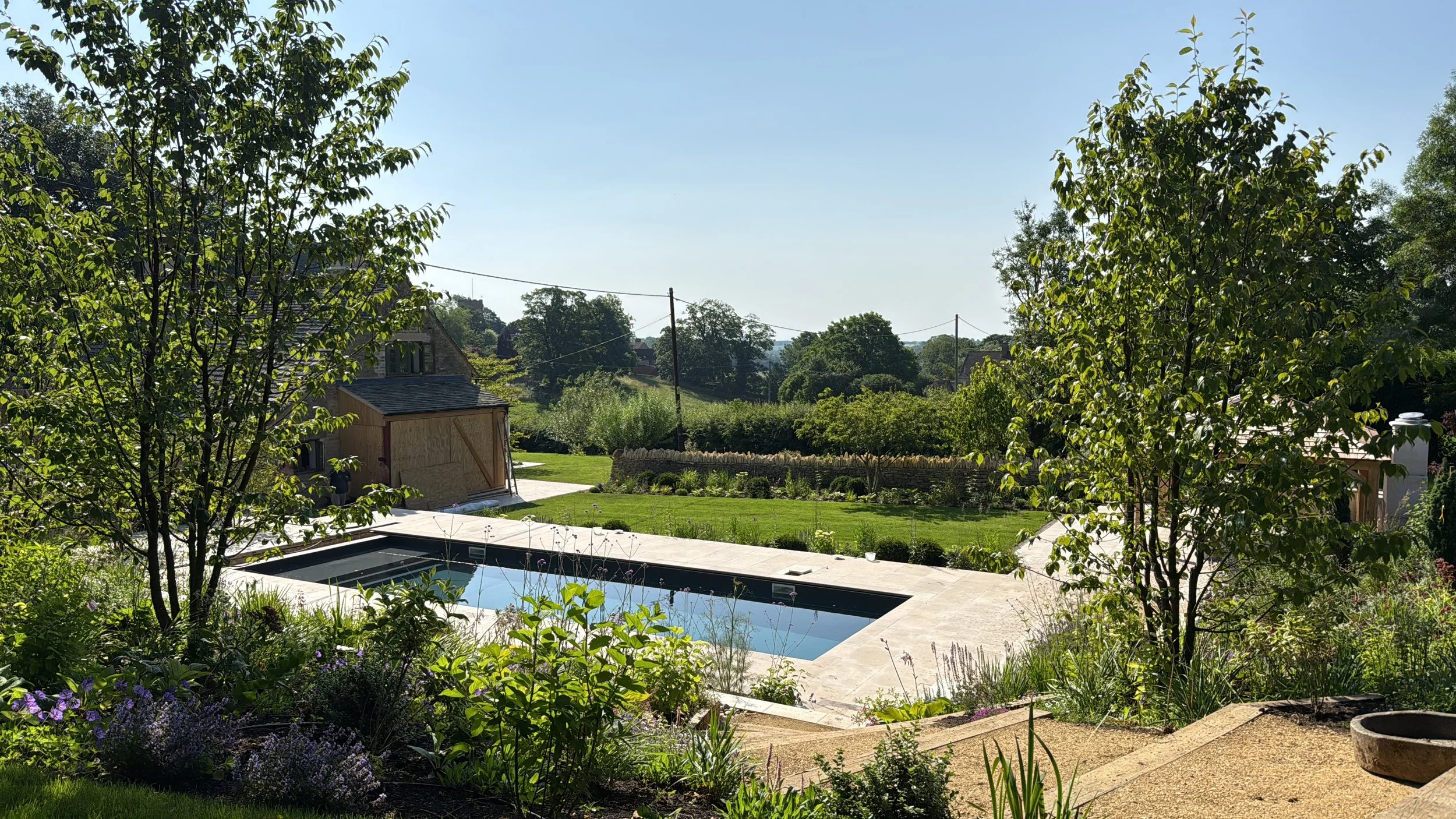 View of a lush garden with trees, a swimming pool, and a picnic area under a clear blue sky.