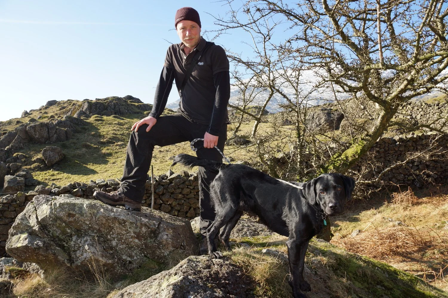 A man with a beard wearing a dark beanie and black outdoor clothing standing on a large rock with a black dog on a leash in a rural, mountainous landscape with rocks, grass, and leafless trees.